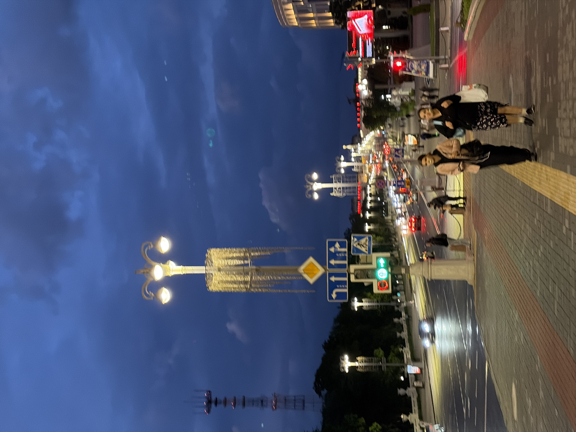 The image depicts a street scene at night in Minsk, Belarus. The sky is a deep blue, suggesting either dusk or night. The foreground features a wide sidewalk paved with gray and red bricks, with a yellow tactile strip for the visually impaired running along its edge. Two people are walking on the sidewalk, a woman in a black dress with floral patterns and a woman in a black outfit with a light pink jacket.
The street is illuminated by streetlights, including a decorative multi-lamp post with ornate detailing. Traffic signals are visible, with a green light indicating the go-ahead for traffic. Various road signs are mounted on a post, indicating directions and pedestrian crossings.
Cars are moving on the road, their headlights causing streaks of light due to motion blur. Buildings are visible in the background, some with lit windows. A tall telecommunications tower is visible in the distance. The environment is urban, with elements of city infrastructure and activity present.