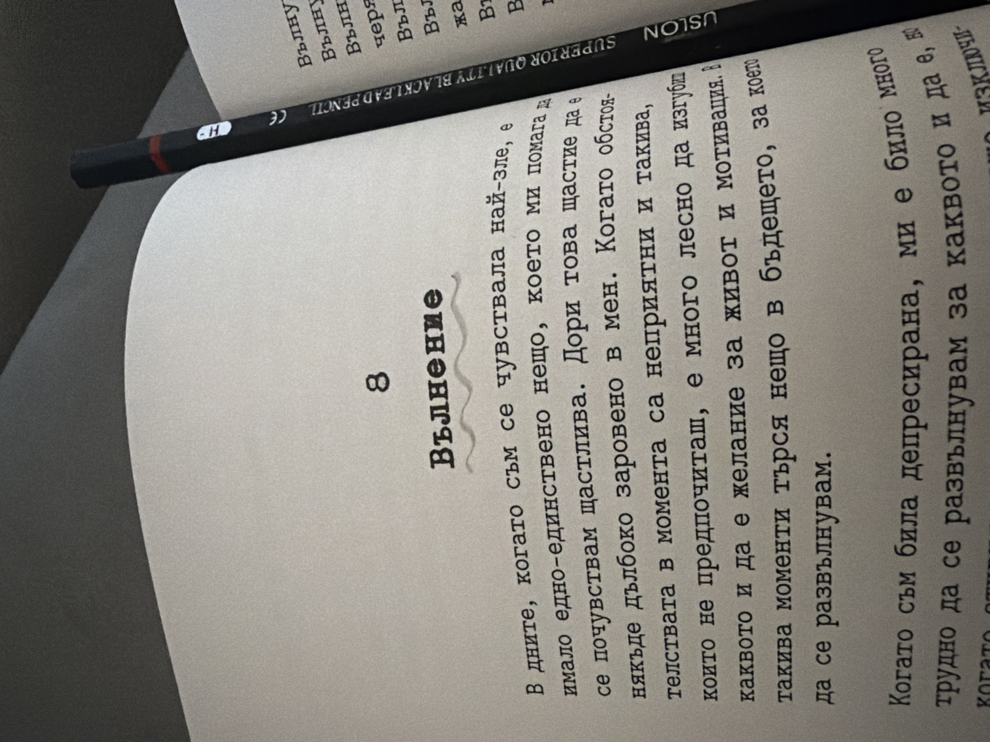 The image shows an open book with text in Bulgarian and a black pencil with "SUPERIOR QUALITY BLACK LEAD PENCIL" printed on it. The book is open to page 8, which has the title "Вълнение" (Excitement). The text on the page discusses feelings of happiness and the desire to live and be motivated, especially during difficult times. There are no people or animals visible in the image. The environment appears to be indoors, with a soft gray surface behind the book, suggesting it might be on a couch or bed. The lighting is dim, casting shadows on the pages. The image does not provide any information about the location in the city of Svilengrad, Bulgaria.