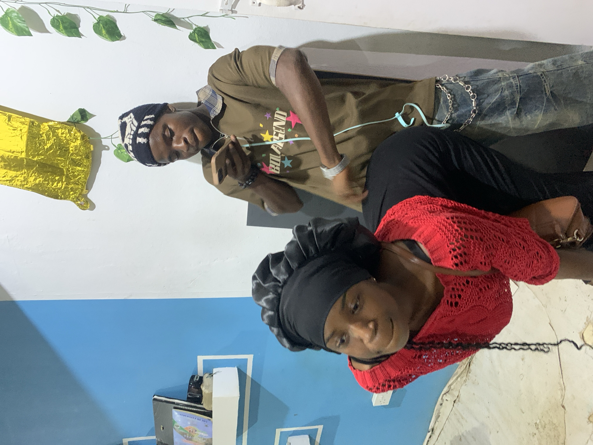 This image shows two young people in an indoor setting, likely a residence in Jos, Nigeria. The background features a blue and white wall, with a shelf on the blue section holding some items, possibly books or binders. A yellow balloon hangs from the ceiling. The person on the left is a young woman wearing a black satin bonnet and a red crocheted top, with her head bowed and looking to the side. The person on the right is a young man wearing a patterned beanie, a brown t-shirt with "BOLAGEND" printed on it, and distressed denim jeans with a silver chain. He is looking at his phone and appears to be interacting with the woman. The lighting suggests it is daytime, though the interior setting makes it difficult to determine the exact time. The floor appears to be concrete or tiled.
