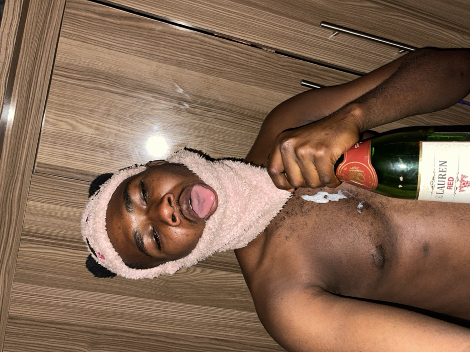 A shirtless male individual with dark skin is depicted, wearing a pink, fuzzy head covering resembling a panda balaclava, which extends over his head, ears, chin, and neck. He holds a green glass bottle labeled "ST. LAUREN RED" with a red cap in his left hand. His tongue is extended from his mouth. A white, viscous substance is visible on his chest, dripping from below the head covering. The background consists of vertical light brown wood-grained cabinetry or paneling, with a reflective horizontal strip visible near the top. The specific location within Muchiya, Nigeria, cannot be determined from the visual evidence provided in the image.