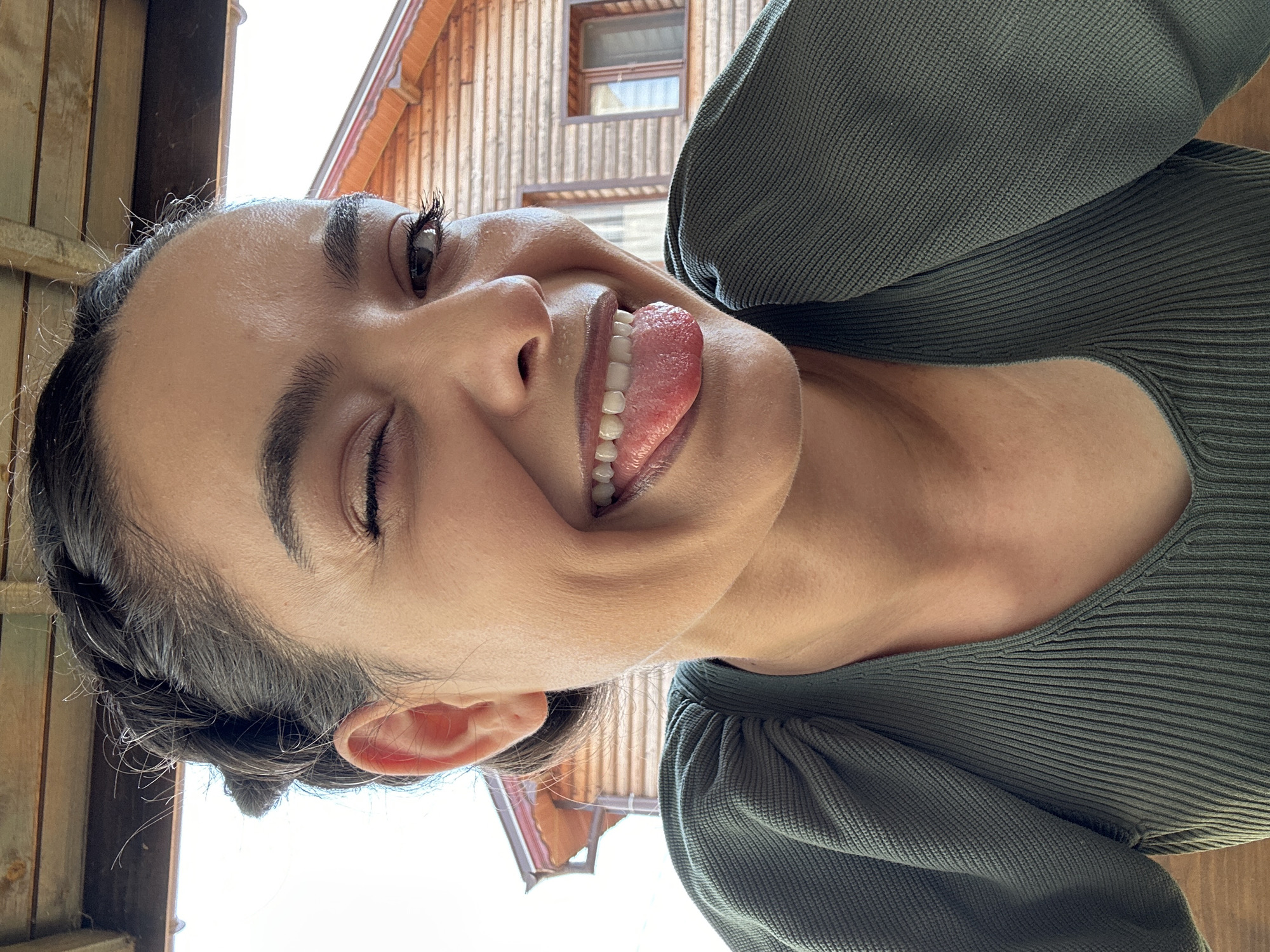 A close-up, selfie-style shot shows a young woman with her tongue sticking out and one eye winking. She has dark hair styled in a braid and is wearing a dark green, ribbed top with puffed sleeves. The background consists of a wooden structure, possibly a building or porch, with exposed beams and a section of a wooden house with a window. The scene appears to be outdoors, under a canopy. There is no indication of the city of Malevo, Bulgaria.