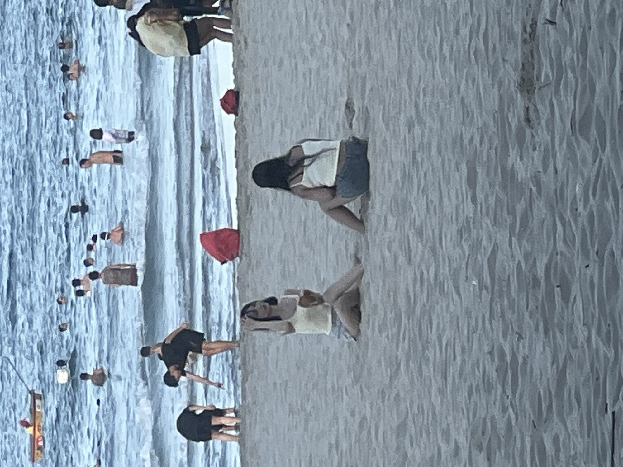 The image depicts a lively beach scene at My Khe Beach in Da Nang, Vietnam, on a cloudy day. The water is a hazy blue, with small waves lapping at the shore. Several people are visible in the water, some swimming and others wading. In the foreground, two young women are sitting on the sand, facing away from the camera. One is wearing a white tank top and denim shorts, while the other has on a yellow tank top and shorts. Both are engaged in personal activities, with one woman touching her hair and the other holding something in her hand. In the background, more beachgoers are scattered across the sand and in the water, some with their backs to the viewer. A red bag is visible near the water's edge. The overall atmosphere is one of relaxed recreation, with a hint of the busy energy of a popular tourist destination.