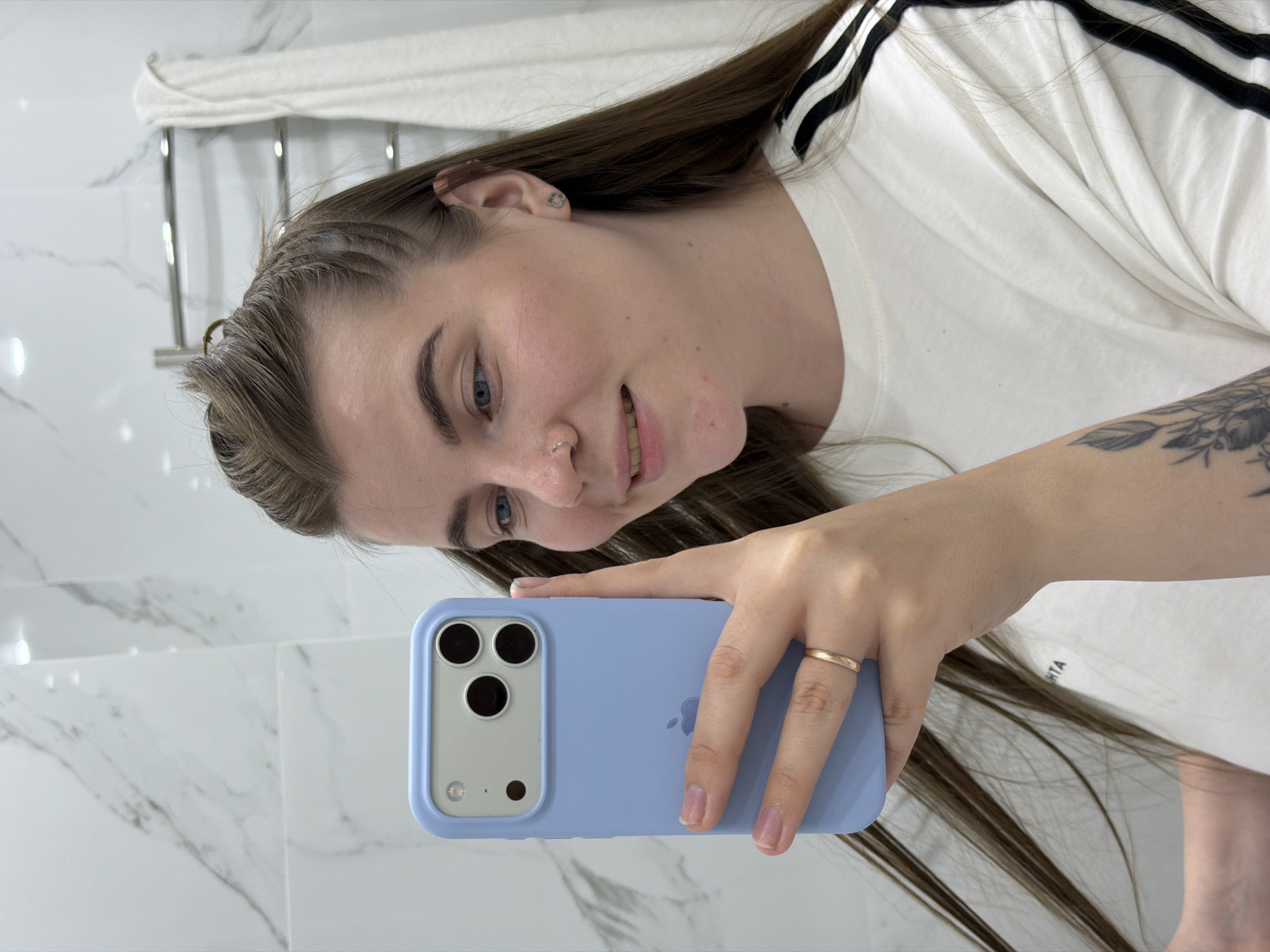 A young woman is taking a selfie in a bathroom. She is holding a blue smartphone with a silver camera module. The phone has three lenses and a flash. She is wearing a white t-shirt with black stripes on the sleeves, and a gold ring on her ring finger. The bathroom has white marble walls and a towel rack with a white towel. Her hair is long and brown, and she has a nose ring. Her left arm shows a tattoo. The image doesn't contain enough information to confirm that it was taken in the city of Chaikovsky, Russia.