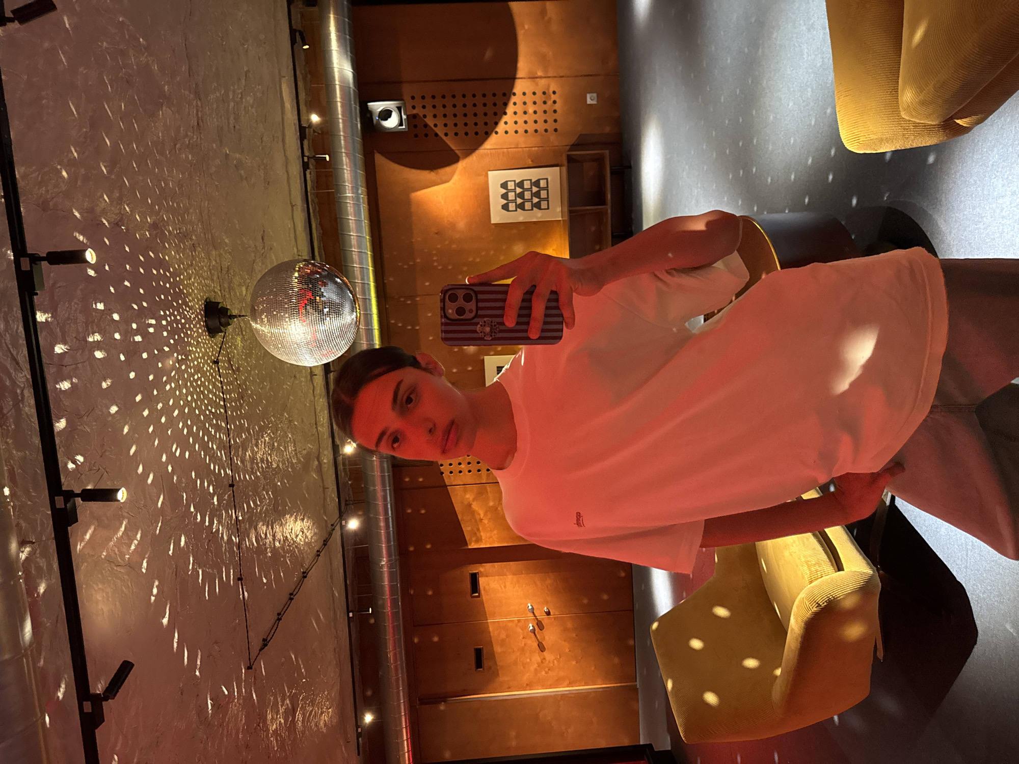A young person is taking a selfie in what appears to be an entertainment venue. The person is wearing a white T-shirt and standing in front of a wall with a disco ball attached. The disco ball is reflecting light onto the wall and creating a pattern. A yellow arm chair is in the right side of the photo. There is artwork visible on a wall behind the person. The image suggests a nightlife setting. There is no discernible evidence to ascertain a location in Yerevan, Armenia.