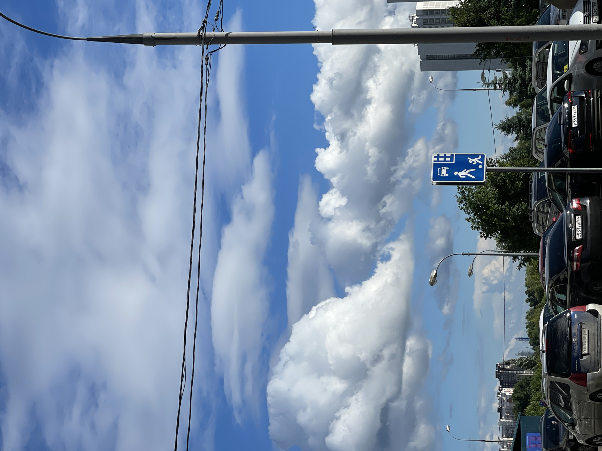 The image depicts a street scene in Moscow, Russia, on a clear day with dramatic clouds. In the foreground, several cars are parked. A blue traffic sign is visible on the right side, indicating pedestrian and children's play areas near a roadway. A tall lamppost stands prominently on the right, with power lines stretching across the upper part of the frame. In the background, apartment buildings and trees can be seen, suggesting an urban or suburban residential area. The sky is a vibrant blue, filled with large, white cumulus clouds. The overall impression is of a typical summer day in a Russian city.