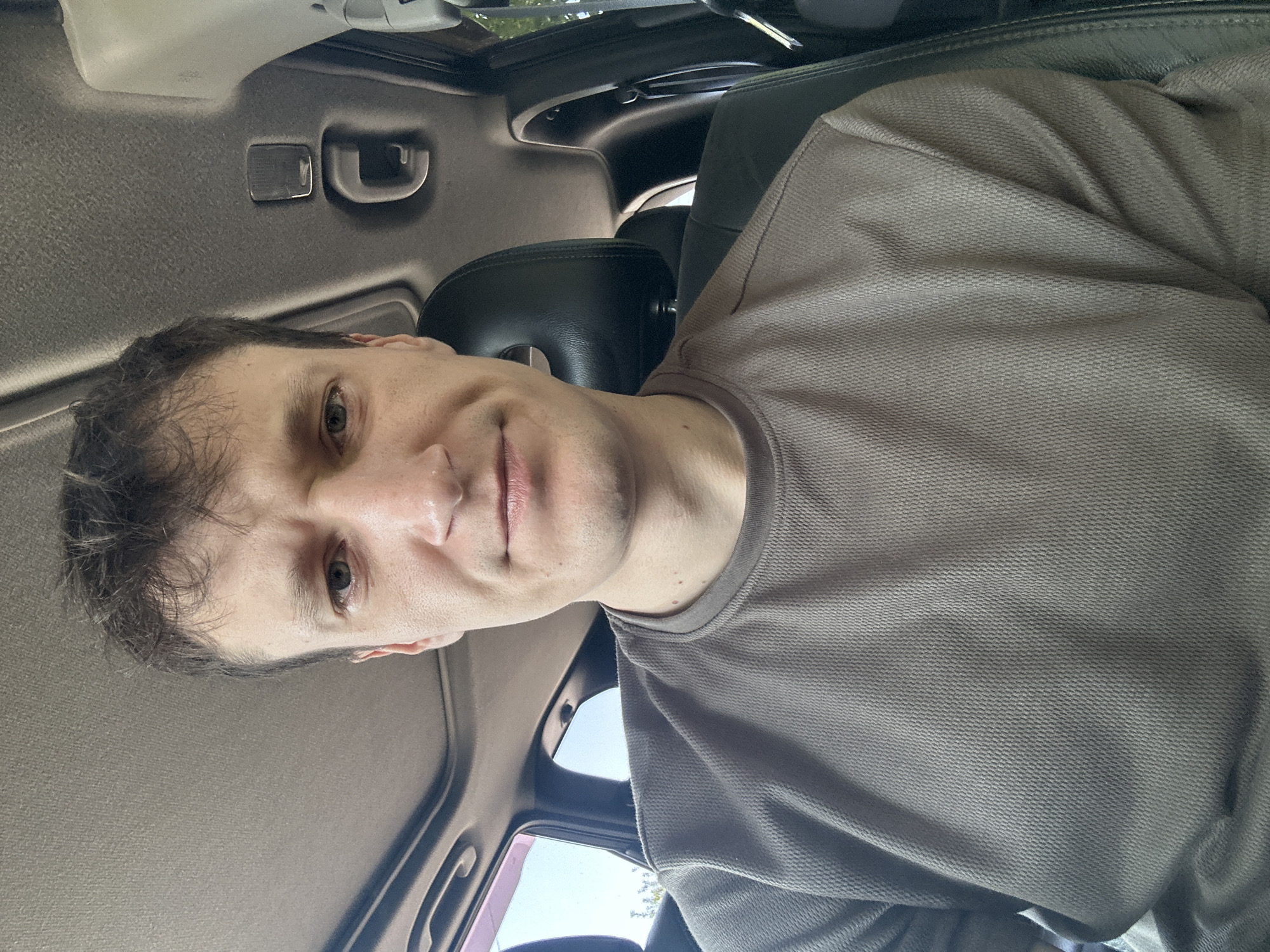 A man with brown hair, green eyes, and a light complexion is sitting in the driver's seat of a car. He is wearing a brown t-shirt. The car's interior is visible, including the headrest, sunroof, and part of the dashboard. The man is looking directly at the camera with a neutral expression. The background suggests an outdoor environment, possibly a street or parking lot, with glimpses of trees and sky through the car windows. The lighting is natural, indicating it is daytime. The image was taken in Makiv, Ukraine.