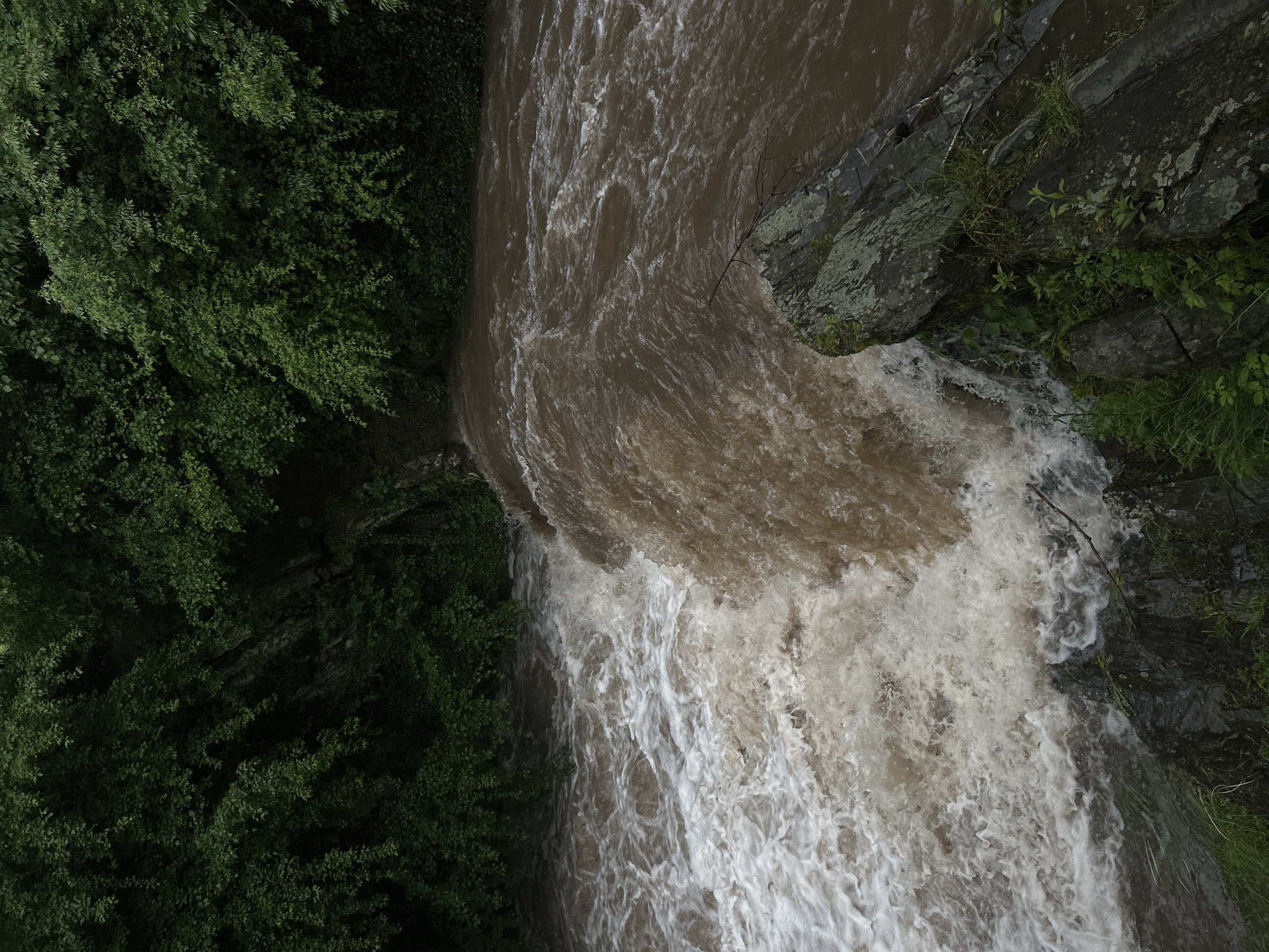 An aerial view depicts a waterfall in the city of Unknown, Unknown. The water is brownish and churning, with white foam where it cascades down. Lush green trees line the left side of the image, while moss-covered rocks are visible on the right.