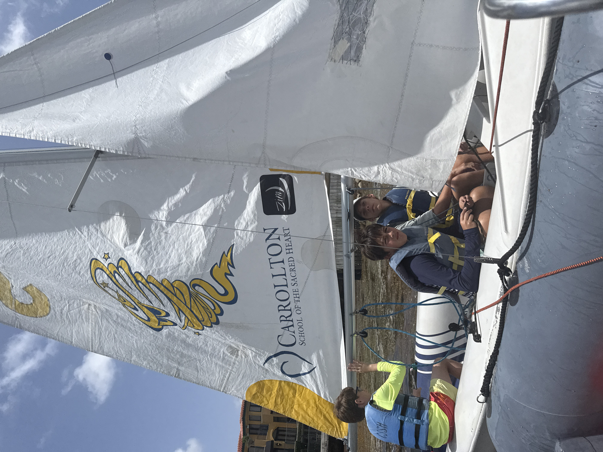 Three boys are in a sailboat on a body of water, with buildings in the background. The sailboat has a white sail with a large yellow and blue logo that reads "CARROLLTON SCHOOL OF THE SACRED HEART". The boy on the left is wearing a blue life vest over a yellow long-sleeved shirt and red shorts. He is looking to the left. The boy in the middle is wearing a blue life vest and a dark blue shirt, and is looking forward. The boy on the right is wearing a blue life vest over a dark blue shirt, and is looking forward. The water appears murky and there is some vegetation floating on the surface. The buildings in the background appear to be apartments or condos, with balconies and tiled roofs. The sky is blue with some white clouds. The sailboat is likely a Zim sailing dinghy, commonly used for training. The Carrollton School of the Sacred Heart is located in Miami, Florida.