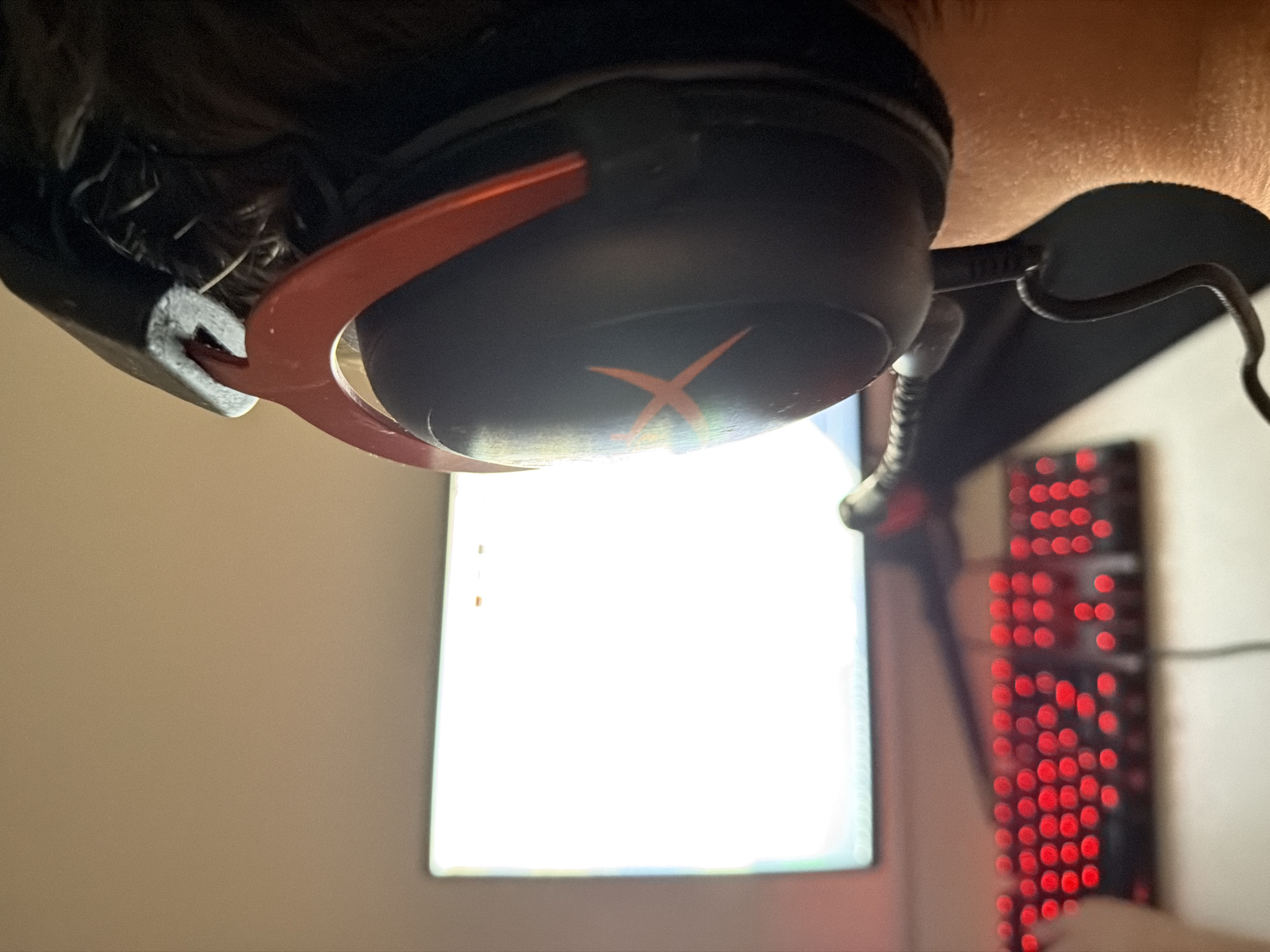 A close-up, slightly upside-down shot depicts a person wearing a black and orange gaming headset. The headset features an orange "X" logo on the earcup. The person's hair is visible around the headset. In the background, a computer monitor displays a bright, mostly white screen. To the right of the monitor, a gaming keyboard with red LED backlighting is partially visible. The location in Budva, Montenegro cannot be determined from the image.