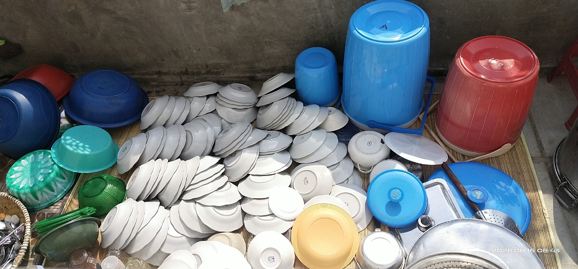 The image depicts an outdoor scene in Muka Haji, Indonesia, where a collection of household items, primarily plastic containers and ceramic dishes, are arranged on a woven mat. The items are placed against a concrete wall.
Prominently featured are several large plastic buckets: two are blue, one is red, and there is a smaller blue container. Adjacent to these are numerous white ceramic plates and bowls stacked in piles. Several smaller plastic basins in green and teal are also visible. Among the dishes, there are also some plastic lids and a metal sieve. A basket filled with spoons and other utensils is partially visible on the left side of the image.
The overall impression is that of items being stored or perhaps prepared for washing or use outdoors. The lighting suggests it is daytime. No people are present in the image.