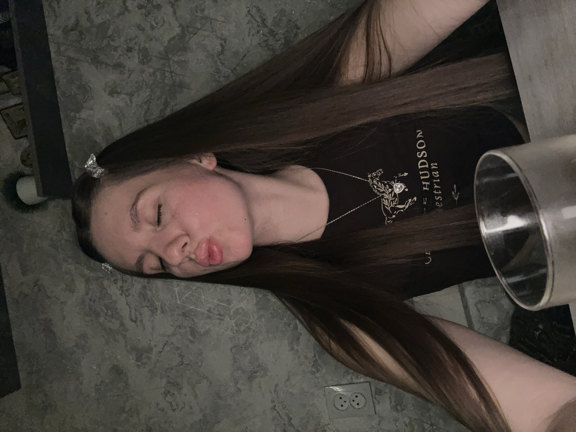 A young woman with long brown hair lies on a gray-patterned surface with electrical outlets visible in the lower center. She has butterfly clips in her hair and is wearing a black t-shirt with the words "Hudson Equestrian." A metallic object is visible on the right side of the frame. The location is not discernible from the image.