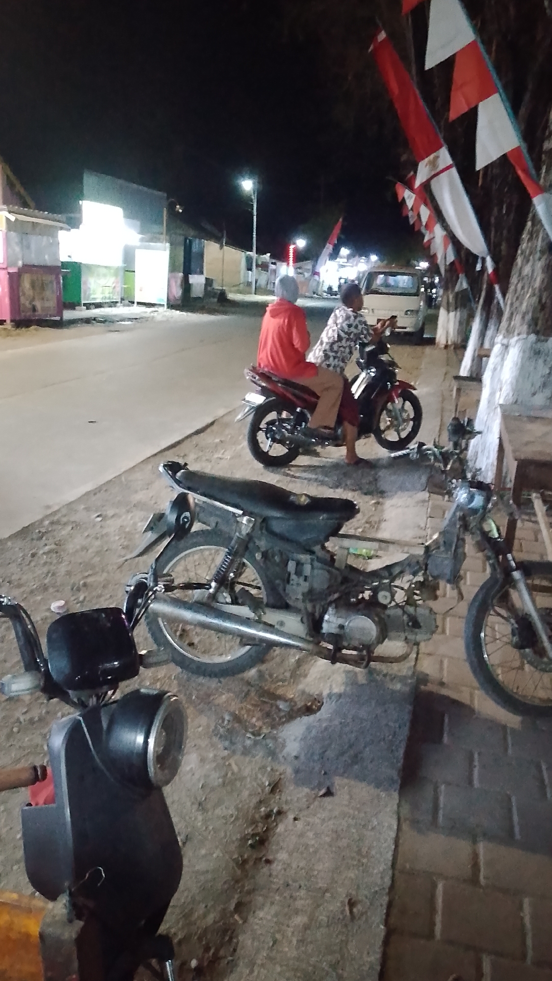 The image depicts a nighttime scene on a street in Karanglangu, Indonesia, likely near a row of small shops. In the foreground, a partially dismantled motorcycle is parked on a dirt and paved surface. To the right, another motorcycle is ridden by two individuals; the rider wears a patterned shirt, and the passenger, in a red hooded jacket, sits behind them. Indonesian flags are visible strung along a tree to the right of the frame. Further back, a white van is parked, and more shops with illuminated signs line the street. The lighting suggests artificial illumination from streetlights and shop displays, casting shadows on the ground.