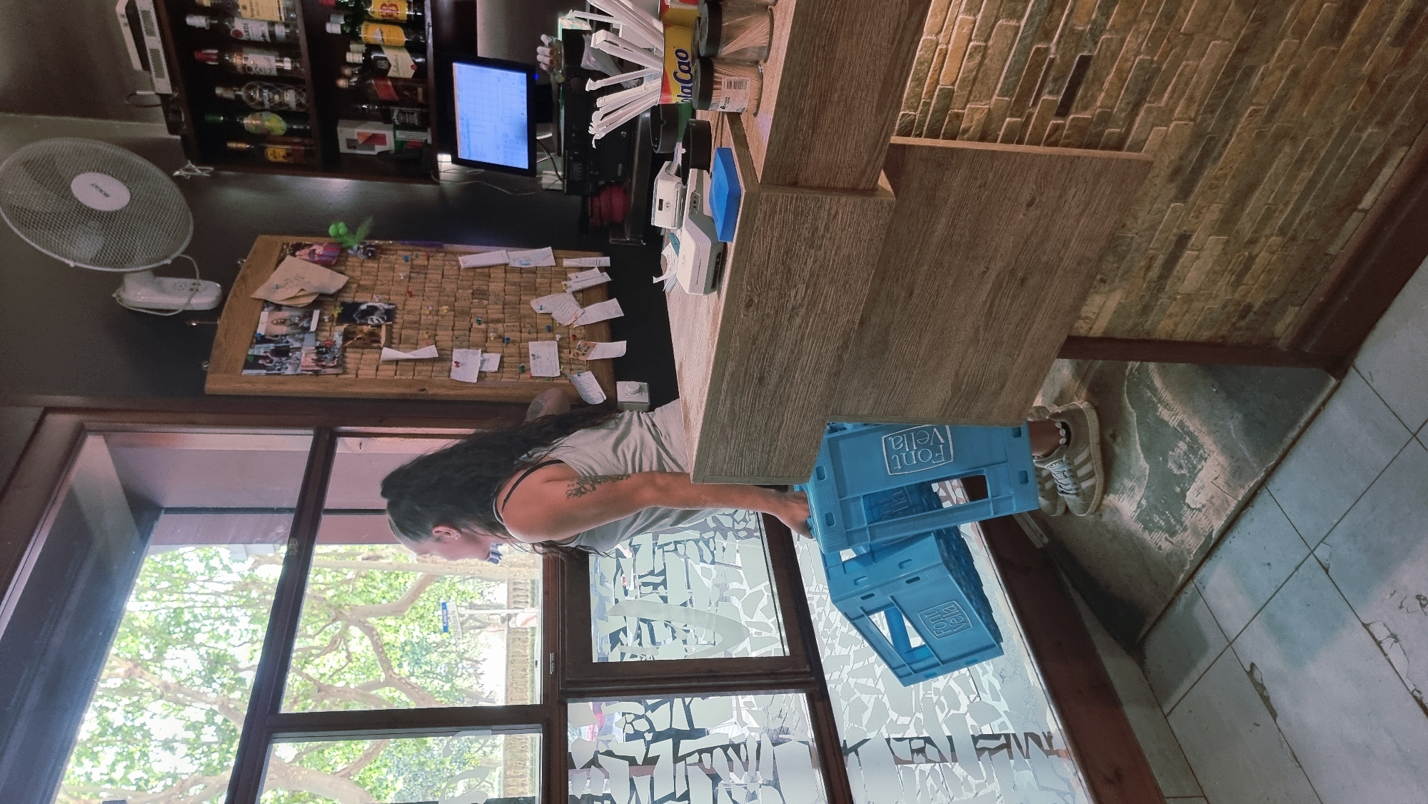 The image shows a woman with long dark hair, wearing a grey tank top and black strap, standing in what appears to be a bar or shop. She has tattoos on her arm. She is reaching for a blue plastic crate with text that appears to say "Font Vella". To her left, there is a large window with a tree branch visible outside. Behind her, there is a corkboard with papers and photos pinned to it. Further back, there are shelves stocked with bottles, a computer monitor, and a desk with various items. The room has a rustic feel with wooden surfaces and a tiled floor. The lighting suggests it is daytime, and the image is taken from a slightly tilted perspective.