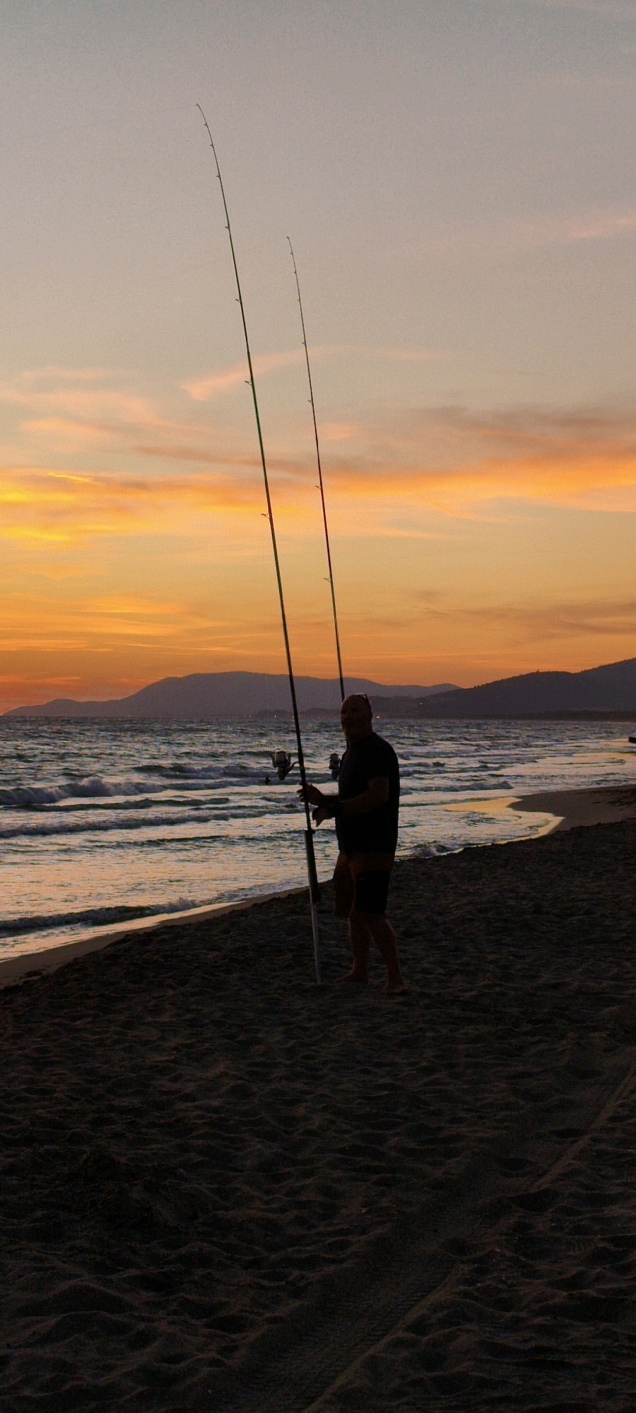 A silhouette of a man fishing on a beach at sunset. The man is standing on the sand, holding two fishing rods. The ocean is to his left, with waves breaking on the shore. The sky is a vibrant orange and yellow, with clouds forming streaks of color. Distant mountains are visible on the horizon. The sand is dark and textured, suggesting it is wet from the tide. The scene appears to be on a beach near Grosseto, Italy.