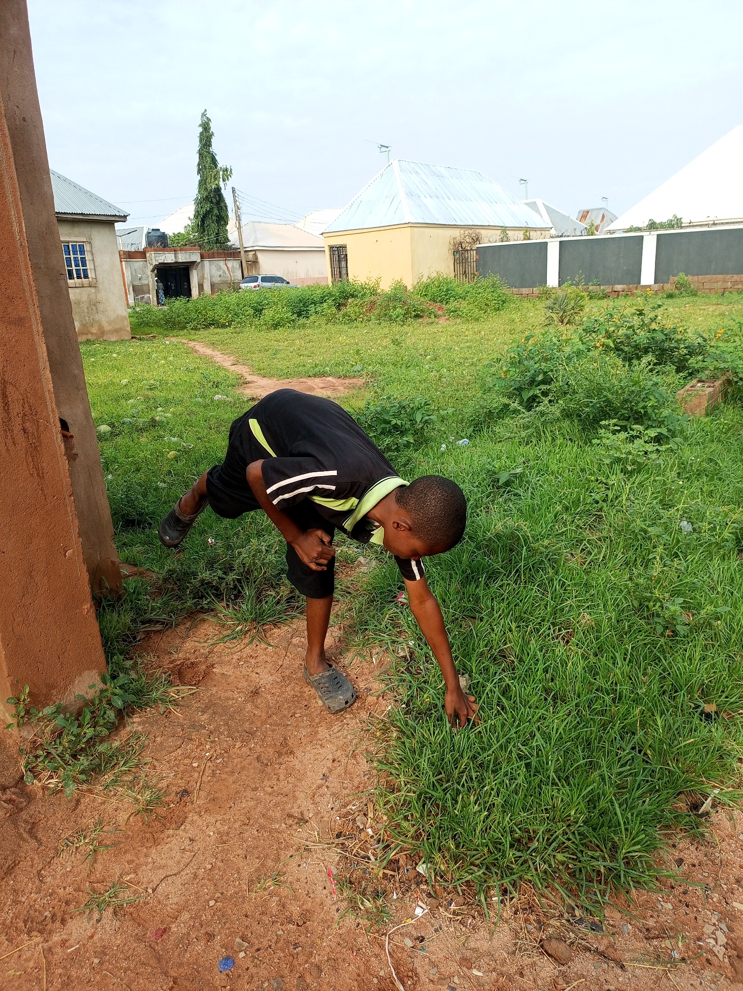 A young boy wearing a black t-shirt with green and white accents and black shorts is bending over to touch the grass in a grassy area with a dirt path. He is standing on one leg, with his other leg bent and extended backward. His face is turned downwards towards the grass. He appears to be near a reddish-brown, rough-textured structure on the left side of the image. In the background, there are several buildings with various colored roofs, a tall green tree, and a wall. The sky is overcast. The location is within the city of Tempure, Nigeria.