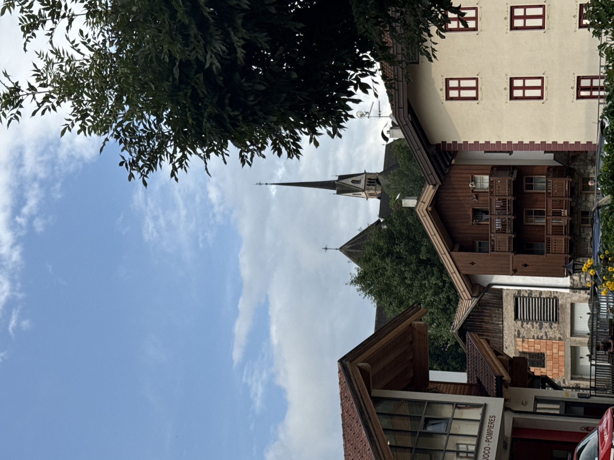 The image captures a street scene in the city of Moena, Italy, showcasing traditional alpine architecture. In the foreground, a row of buildings with wooden balconies and tiled roofs is visible. The building on the right is cream-colored with several red-framed windows. To its left, a narrower building with dark wood siding and multiple balconies is present. Further left, a building with a modern facade featuring large windows and the text "JOCO - POMPIERI" is seen, along with a red vehicle. In the background, a tall church steeple with a cross on top rises above the trees, characteristic of the region. The sky is partly cloudy. A few people are visible in the scene, one sitting near a fence and another standing behind a bush. Yellow flowers are blooming in the lower right corner.
