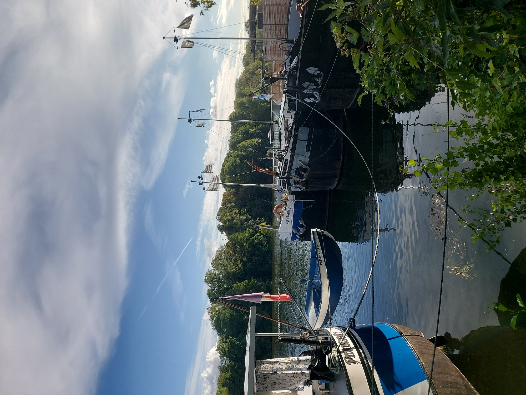 A fleet of dark, industrial barges is moored on a calm river, with a line of trees forming a lush green backdrop along the opposite bank. The sky above is a mix of blue and scattered white clouds, with a faint contrail visible. In the foreground, a smaller blue and white dinghy, named "PARANZELLA," is suspended by a davit from a larger vessel, displaying a French flag. Another barge, labeled "NORMANDIE," is docked to the right, its hull a dark blue and black. The scene appears to be a serene waterside setting, possibly a quay or harbor in Conflans-Sainte-Honorine, France, known for its historical significance as a river port. No people are visible in the image.