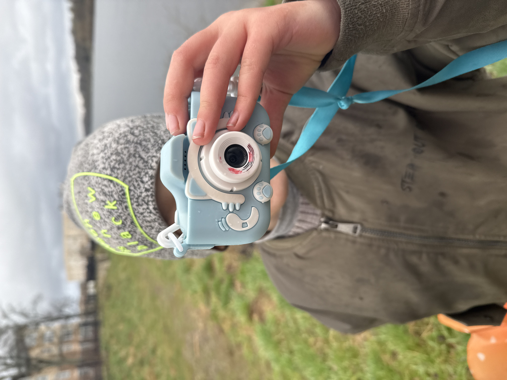 A child holds a light blue toy camera with a white lens and an attached turquoise ribbon. The child wears a grey and white speckled beanie with a neon green trim and letters. They are wearing a grey sweater and an olive green jacket. The background shows a body of water, possibly a river, with trees and foliage along the banks. There is grass visible in the foreground. While the location of the city is indicated in the prompt, there is no way to verify that information.