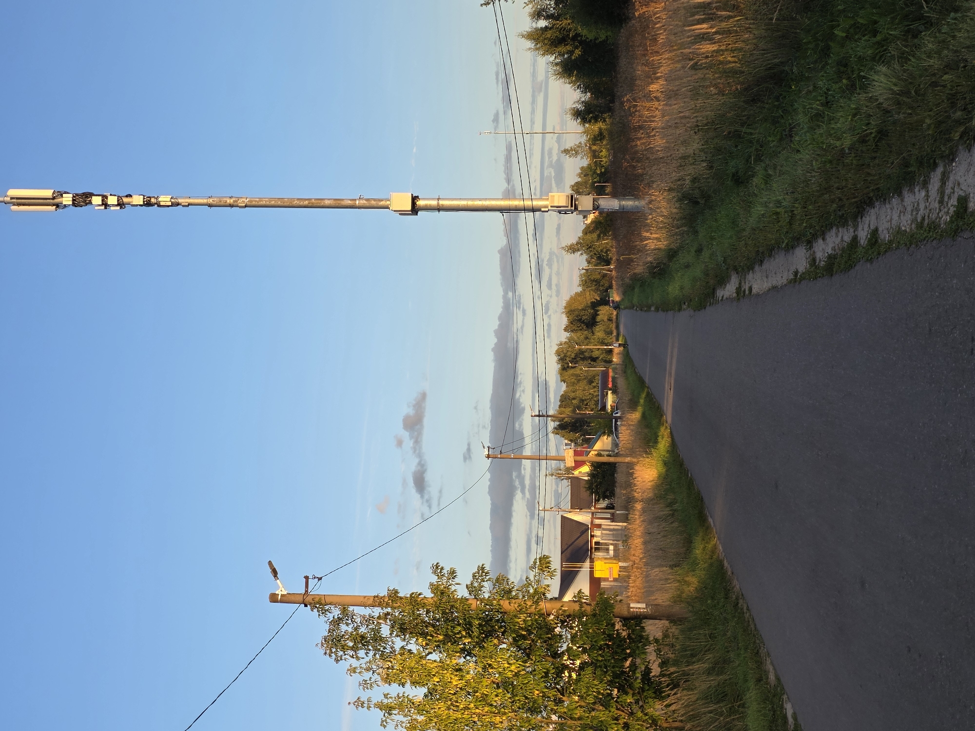 The image depicts a rural road with vegetation on both sides and a clear blue sky. A tall cellular tower with antennas is prominently visible on the right side of the road. On the left side, there is a utility pole with a light fixture and a tree with green leaves. Further down the road on the left, several houses with dark roofs and white walls are situated behind a fence. The sun is casting a warm glow on the scene, particularly on the dry grass and the side of the road. The road appears to be paved and in good condition, with no visible traffic. The overall impression is a peaceful, rural landscape on a sunny day. The location is identified as Rechitsy, Russia.