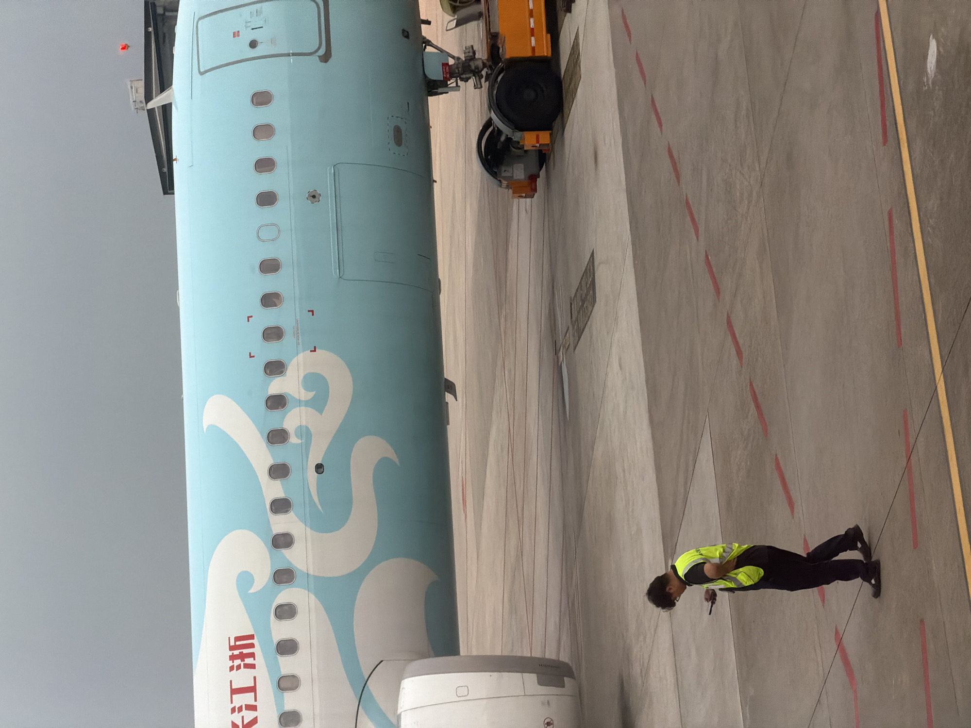 The image depicts the side of an aircraft with a light blue and white livery featuring wave-like designs, along with text in Chinese characters. An aircraft tug is partially visible near the nose of the plane. A ground worker, wearing a high-visibility yellow vest and dark trousers, stands on the concrete tarmac next to the aircraft. Red and yellow lines mark the tarmac surface, potentially indicating designated pathways or zones for aircraft operations. The setting appears to be an airport apron, most likely located in Baiyun District, China, as evidenced by the aircraft's livery which might indicate it is a Zhejiang Loong Airlines aircraft, a Chinese carrier. The sky is overcast, suggesting possible inclement weather.