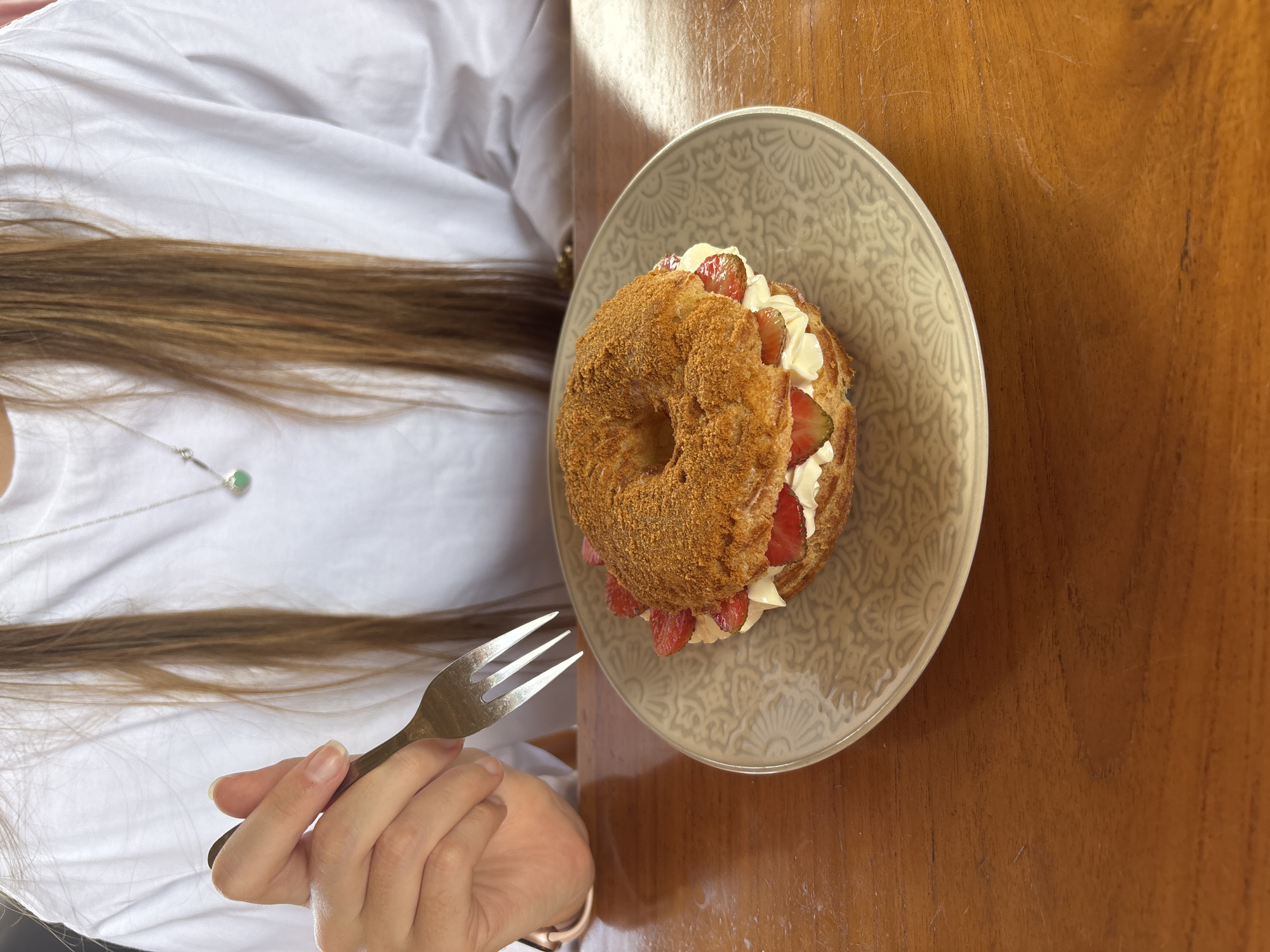 A person's hands are visible, holding a fork over a round pastry filled with cream and strawberries, which sits on a decorative plate. The pastry appears to be a choux pastry or similar, with a golden-brown exterior and a hole in the center. It is cut in half horizontally and filled with white cream and sliced strawberries. The plate is light gray with an intricate, embossed pattern. The pastry and plate are on a wooden table. The person is wearing a white t-shirt and a silver necklace with a green pendant. Long, light brown hair is visible falling over their shoulders. The lighting suggests an indoor setting, possibly a cafe or restaurant, with natural light coming from the side.