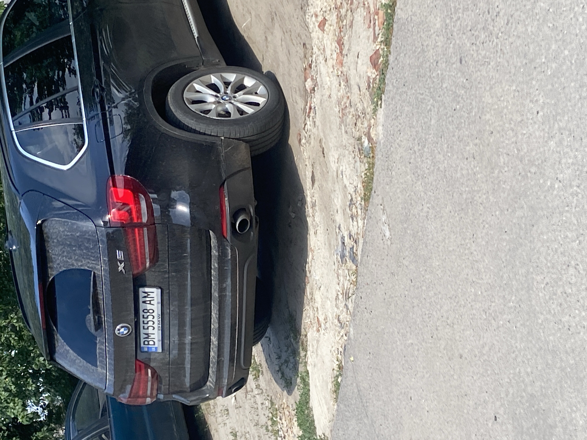 A black BMW X5 is parked on a dusty patch of ground, possibly a parking lot or roadside in Trostianets, Ukraine. The vehicle is oriented with its rear facing the camera. The license plate reads "BM 5558 AM." The car is slightly dirty, with dust accumulated on its surfaces. In the background, there are green trees and foliage, suggesting an outdoor setting. The foreground consists of a paved surface, likely a road or sidewalk, with some scattered debris. No people are visible in the image.