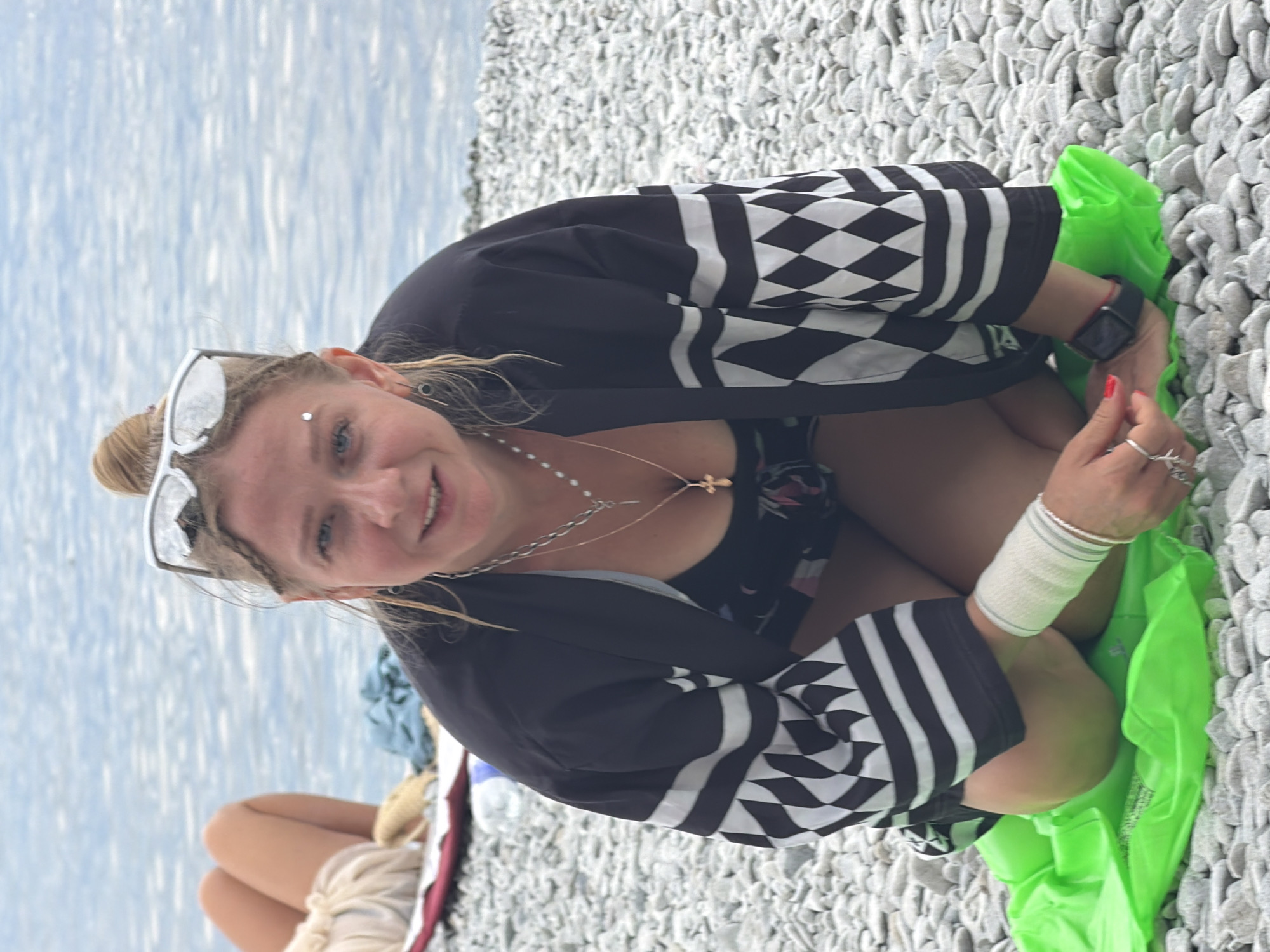 A woman with blonde hair in a bun is kneeling on a bright green inflatable mat on a pebble beach. She is wearing a black and white patterned jacket over a dark swimsuit, a silver necklace with a cross pendant, and a smartwatch. Her left arm is bandaged. She is looking at the camera and smiling. The water is in the background. To her left, a person's legs are visible, also on the beach. The scene is located in Partenit, Ukraine.