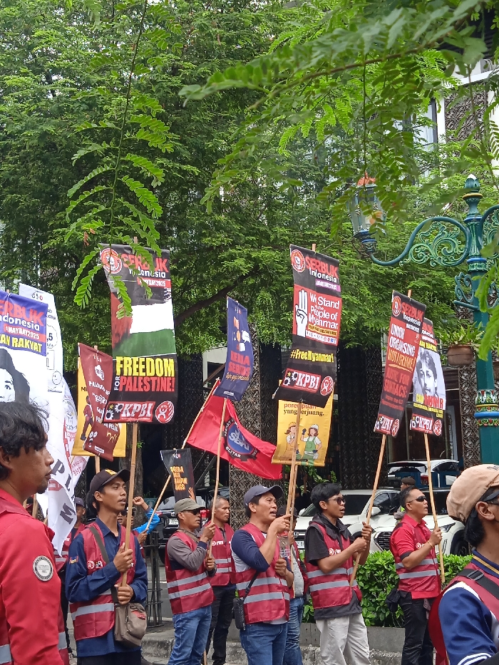 A group of protesters are marching down a street in Yogyakarta, Indonesia (latitude: -7.784, longitude: 110.367), holding signs with slogans in Indonesian and English. The signs advocate for freedom for Palestine, support for the people of Myanmar, and criticize the Indonesian government. Some protesters are wearing vests with the letters "KPBJ" on them. The street is lined with trees, and a street light is visible in the background. The protesters are walking in a single file line, some holding flags. The protesters are diverse in age and gender, but most are male.