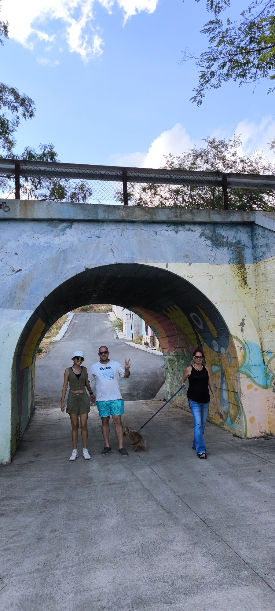 In Fanzara, Spain, under a vibrantly painted arched underpass, three individuals and a small dog are captured in a moment. A woman on the left, wearing a green two-piece outfit and a white bucket hat, stands beside a man in a white t-shirt and blue patterned shorts. He gestures with a peace sign. To the right, a woman in a black tank top and blue jeans walks a small, light-brown dog on a leash. The underpass walls are adorned with colorful graffiti, including rainbow hues and abstract designs. The road ahead is paved and slopes slightly uphill. Above the underpass, a metal railing and chain-link fence separate the structure from trees and a blue sky with scattered clouds.