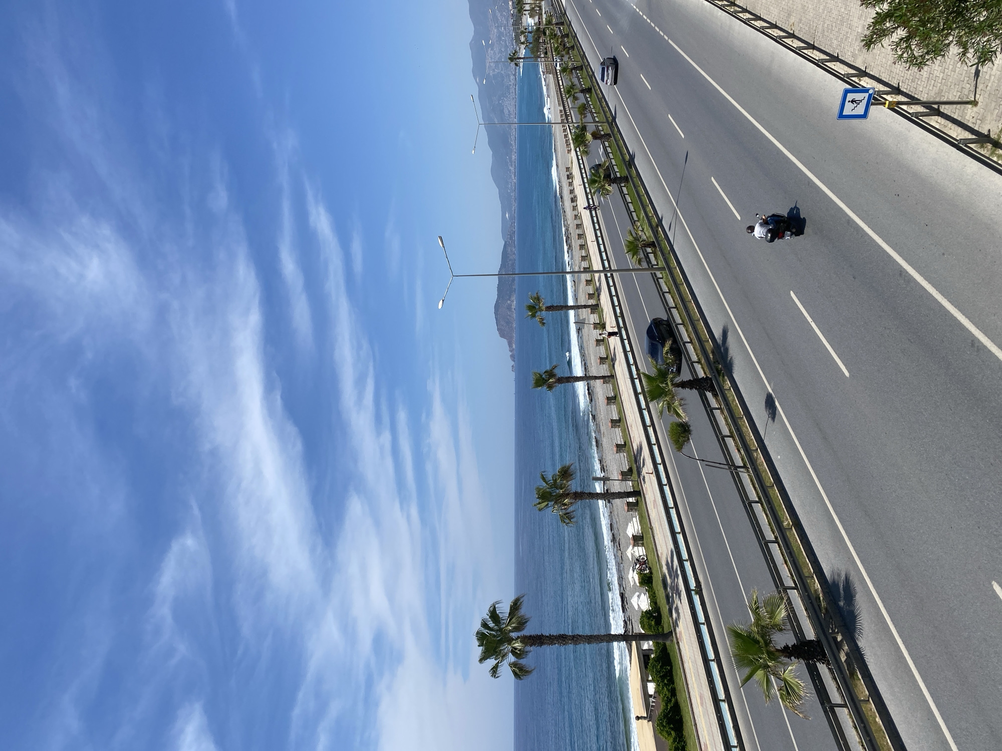 The image shows a coastal road in Alanya, Turkey, with the Mediterranean Sea to the left and mountains in the background. A multi-lane road runs parallel to the coastline, featuring light traffic including a motorcycle. Palm trees line the divider between the road and the beach. The sky is blue with sparse clouds. A beach with some people and white sun umbrellas are visible. A blue road sign indicates the presence of a staircase.