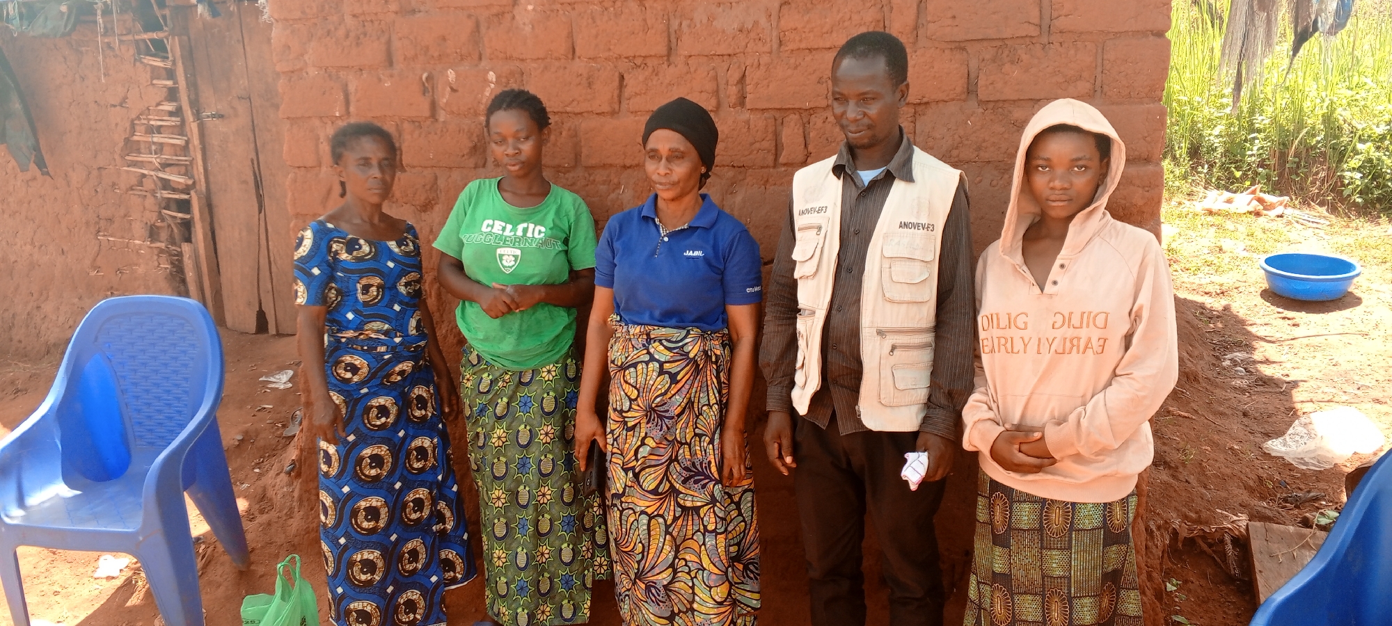The image depicts a group of five individuals standing outdoors in what appears to be a rural setting in Makungu, Democratic Republic of the Congo. Behind them is a reddish-brown brick or mud-brick wall of a building, with an adjacent dilapidated structure.
From left to right:
1. A woman wearing a blue patterned dress and carrying a green plastic bag.
2. A woman wearing a green "Celtic Jugglernaut" t-shirt and a patterned skirt.
3. A woman wearing a blue polo shirt with a "JABIL" logo, a black head covering, and a patterned skirt.
4. A man wearing a brown button-down shirt, a beige vest with pockets and "ANOVEV-E3" printed on the front. He is holding a white object (possibly a cloth) in his hand.
5. A young person wearing a light-colored hooded sweatshirt with the words "DILIG DUIG EARLY LYJAAJ" printed on the front, and a patterned skirt.
The setting appears to be a yard or common area. To the left is a blue plastic chair, and to the right, a blue basin on the ground. The background shows green vegetation. The lighting suggests it is daytime. The individuals are generally looking towards the camera.