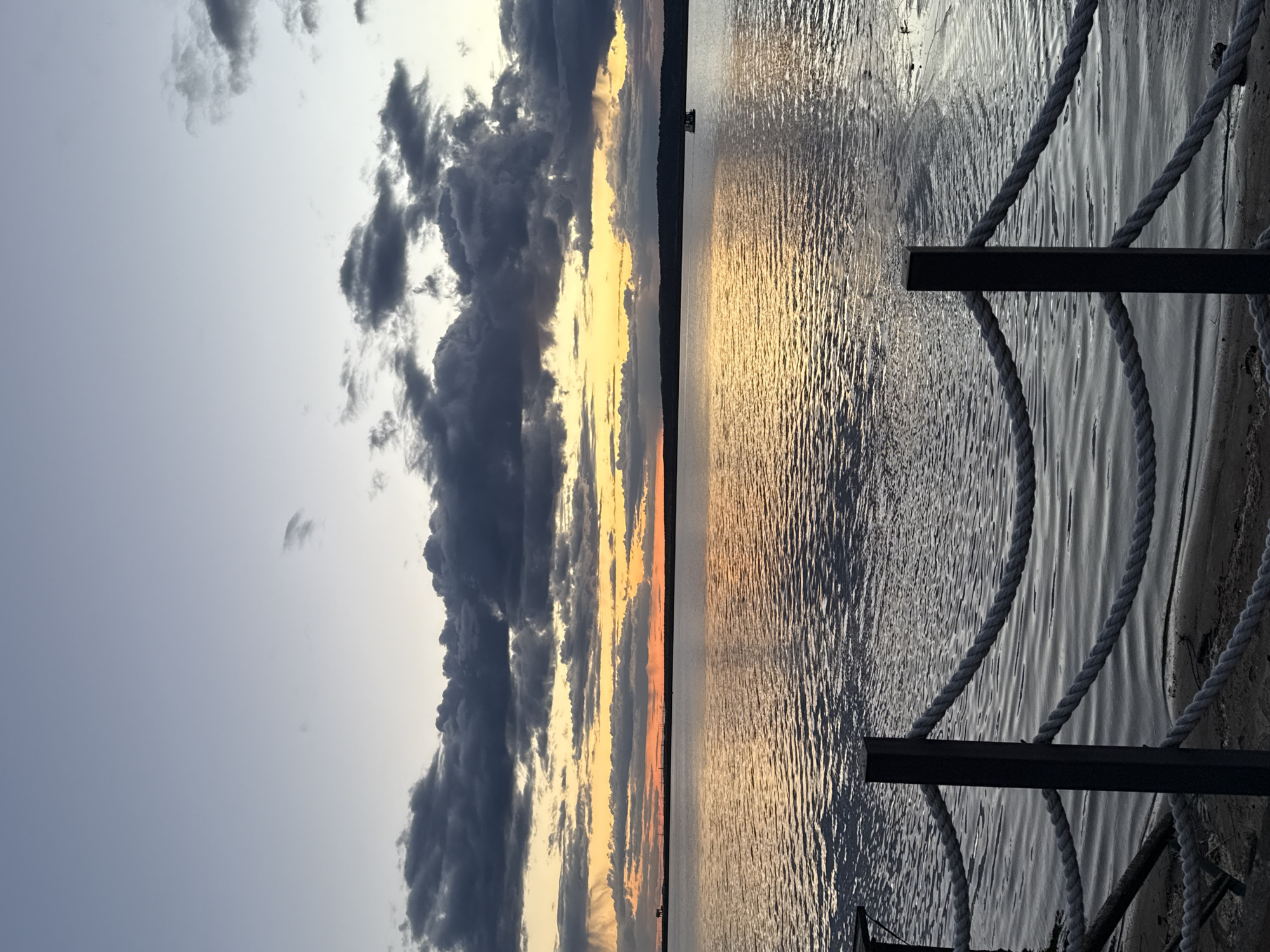 The image captures a waterscape at sunset. In the sky, dark, textured clouds contrast with the lighter, golden hues of the setting sun. Below, the water reflects the colors of the sky, creating a rippled and shimmering surface. A partially visible pier structure with ropes is situated on the right side of the frame. The location is the city of Chaikovsky, Russia.