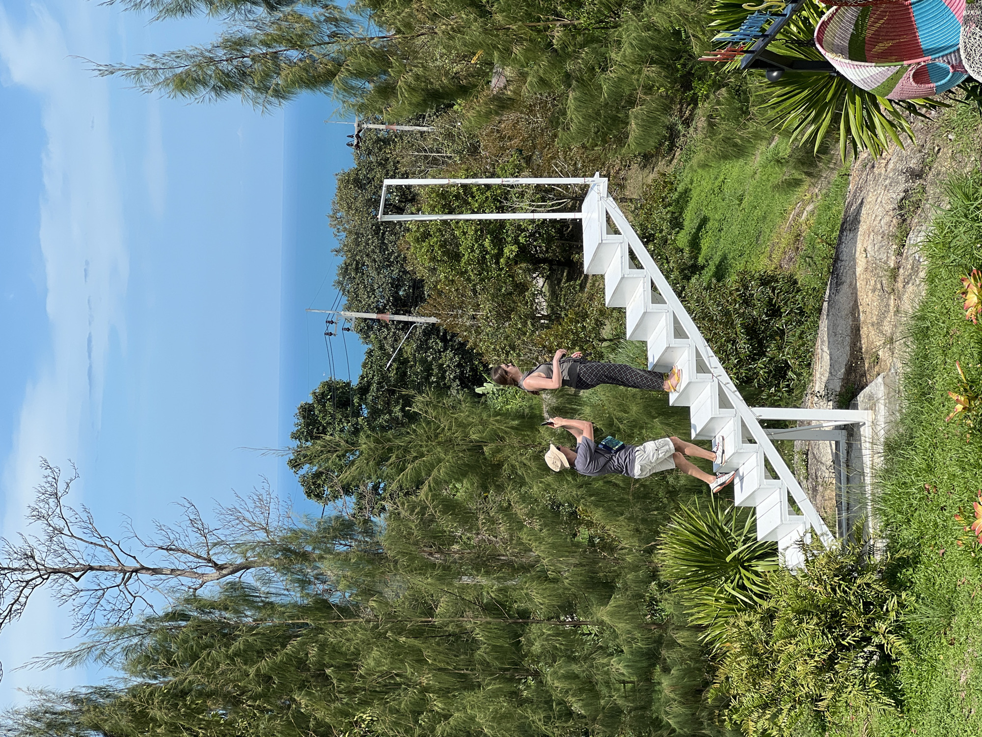 This image captures two individuals on a white sculptural staircase overlooking a distant ocean vista in Baan Thurian, Thailand. On the third step from the bottom, a male with fair skin, wearing a beige bucket hat, grey t-shirt, khaki shorts, and white sneakers, holds a black smartphone horizontally, aimed forward. A blue and white shoulder bag is visible across his chest. Ahead of him, on the top step, a female with fair skin, dark hair tied back, wearing a dark tank top, patterned dark trousers, and yellow flip-flops, stands near a white, open-frame archway. She faces right, looking towards the ocean and sky. The white staircase ascends from left to right, featuring five visible steps and a supporting frame. The scene is surrounded by dense green vegetation, including tall, slender evergreen trees flanking both sides and various shrubs and grass in the mid-ground and foreground. In the background, a clear blue sky with a few faint clouds transitions to a calm, deep blue ocean at the horizon. Utility poles and power lines are visible in the mid-ground, positioned above some of the trees. The lower right foreground includes a colorful, striped woven basket attached to a dark pole with a partially visible blue sign. The environment is brightly lit by natural daylight.
