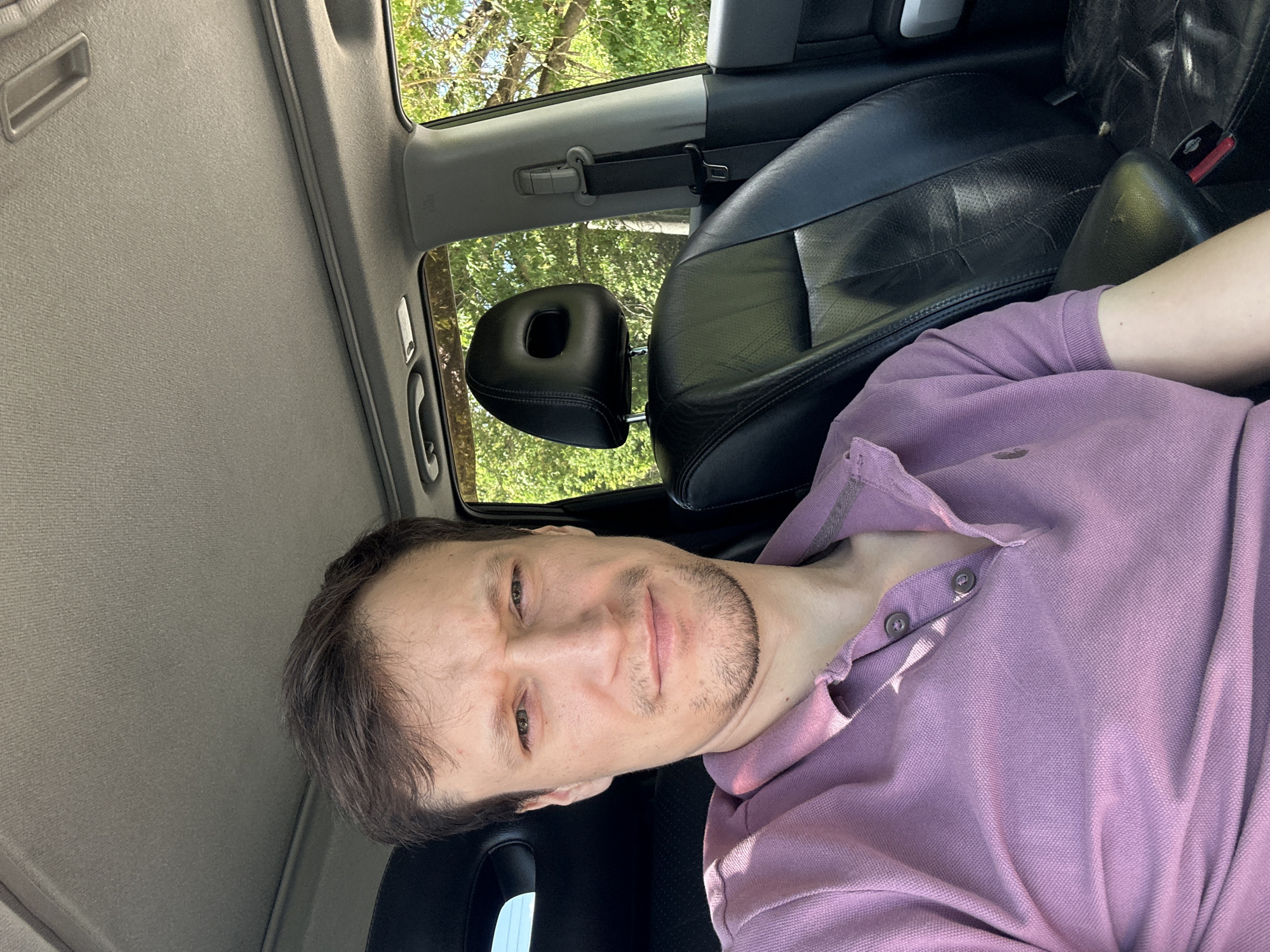 A man with brown hair and light skin is taking a selfie inside a car. He is wearing a purple polo shirt. The interior of the car is visible, including black leather seats and the ceiling. Outside the car, trees and foliage are visible, suggesting an outdoor location. The image appears to be taken during daylight. The man's facial expression is neutral. The location is the city of Makiv, Ukraine.