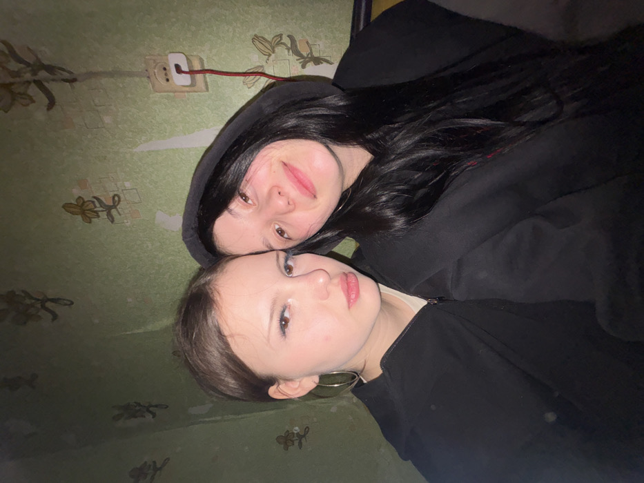 Two young females are depicted in a close-up indoor photograph taken in Foki, Russia. The female on the left has light brown hair, fair skin, and wears large silver hoop earrings and a black zippered top over a light-colored undershirt. She maintains a neutral expression with slightly parted, pink lips and visible blue-tinted eyelashes. The female on the right has long dark hair, which frames her face, and wears a black hooded sweatshirt with the hood up. She displays a soft smile. Both subjects are positioned with their heads close together, appearing to be shoulder-to-shoulder.
The background consists of a wall covered in light green wallpaper featuring a repeating pattern of stylized light brown or yellow flowers and geometric shapes. The wallpaper exhibits signs of wear and age, including peeling sections revealing lighter material underneath. A white, rectangular electrical outlet is visible on the upper right side of the wall, with a red cable plugged into its lower socket, extending downwards. The lighting suggests an artificial source, possibly a camera flash, illuminating the subjects prominently against the somewhat dim background.