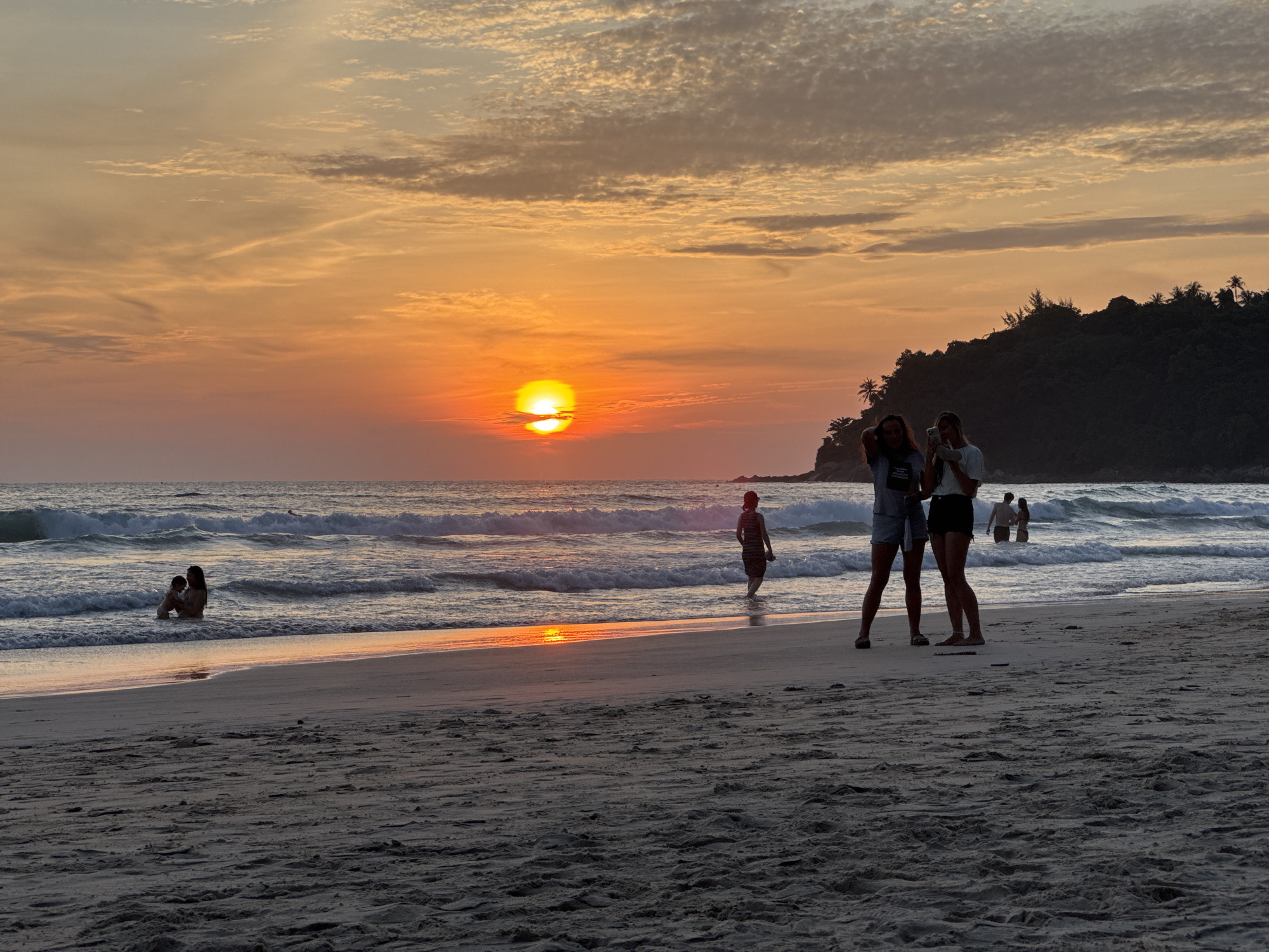 This is a photograph of a beach at sunset in Karon, Thailand. The sky is filled with warm orange and yellow hues, with some scattered clouds. The sun is low on the horizon, casting a golden reflection on the ocean waves.
In the foreground, two young women are standing on the sand, taking a selfie with a smartphone. They are dressed casually in shorts and t-shirts. Further down the beach, a lone figure is walking into the water, and another person is sitting in the shallows with a child. In the distance, a forested hillside is silhouetted against the colorful sky. The beach is sandy, with some debris scattered on the surface. The overall atmosphere is peaceful and serene.