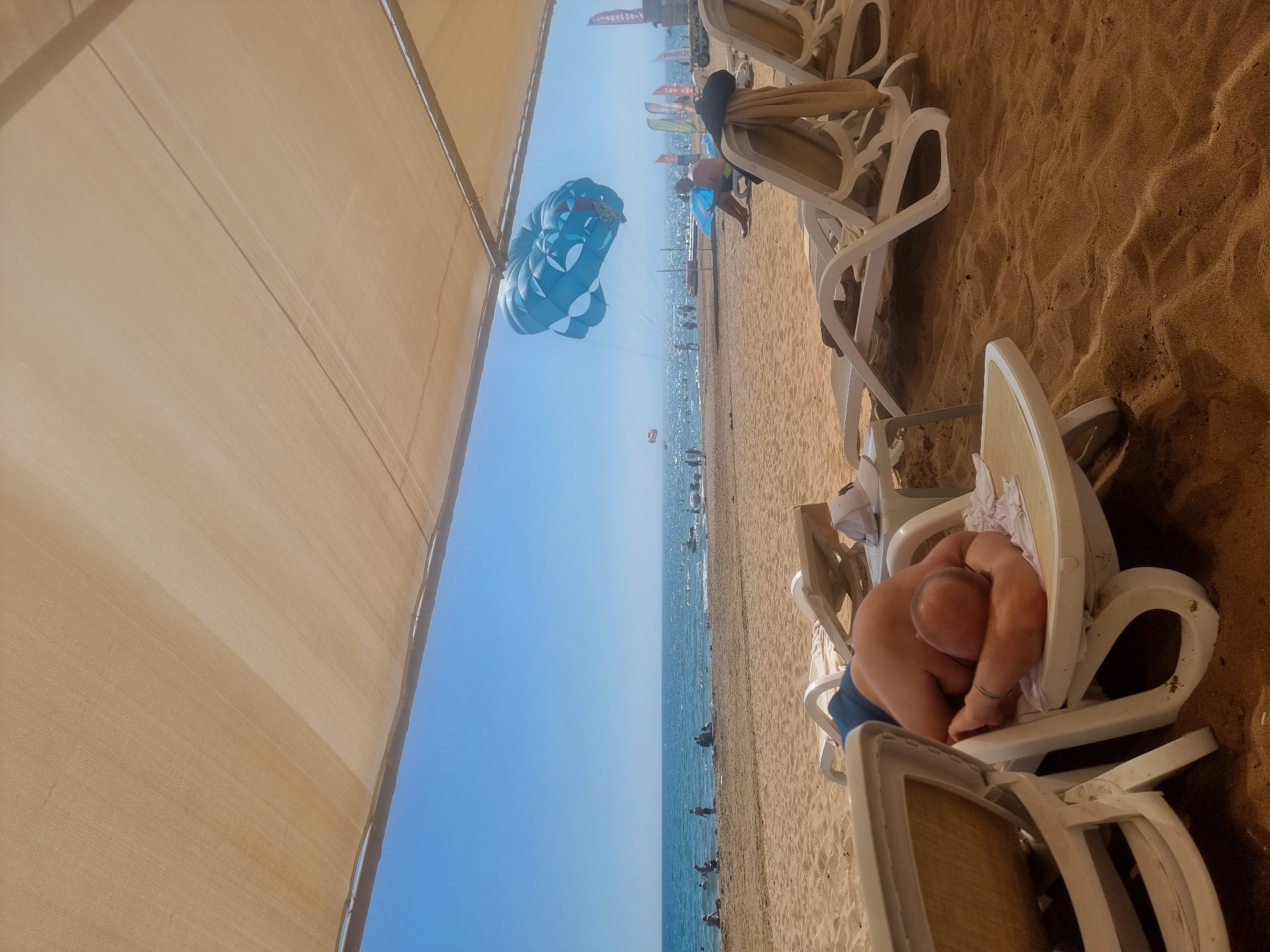 This media file captures a sunny day at a beach in Manavgat, Turkey. The scene is dominated by a large, tan canopy providing shade over a sandy area where white plastic lounge chairs are arranged. In the foreground, a person is lying down on a lounge chair, curled up and seemingly asleep, wearing blue shorts.
Further down the beach, the turquoise ocean stretches out to the horizon under a clear blue sky, suggesting fair weather. Several people are visible in the water, some appearing to be swimming or wading. A prominent blue and white parachute is in the air, indicating parasailing activities are taking place.
In the background, there are more lounge chairs and beachgoers. Some flags and what appear to be signs or banners are visible along the beachfront, adding to the lively atmosphere of a resort or tourist area. The time of day appears to be daytime, likely late morning or early afternoon given the bright sunlight and long shadows cast by the canopy and chairs. The overall emotion conveyed is one of relaxation and enjoyment of a beach vacation.