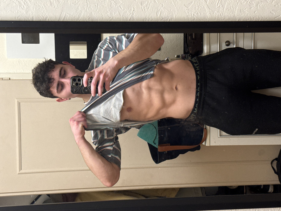 A shirtless man with abs poses in a mirror. He wears black sweatpants and a striped shirt partially pulled up. He holds a phone with a topographical map case, likely taking a selfie. The mirror reflects a partially visible room, including a door and what appears to be a portion of a dresser or cabinet. The photo is taken indoors with artificial lighting.