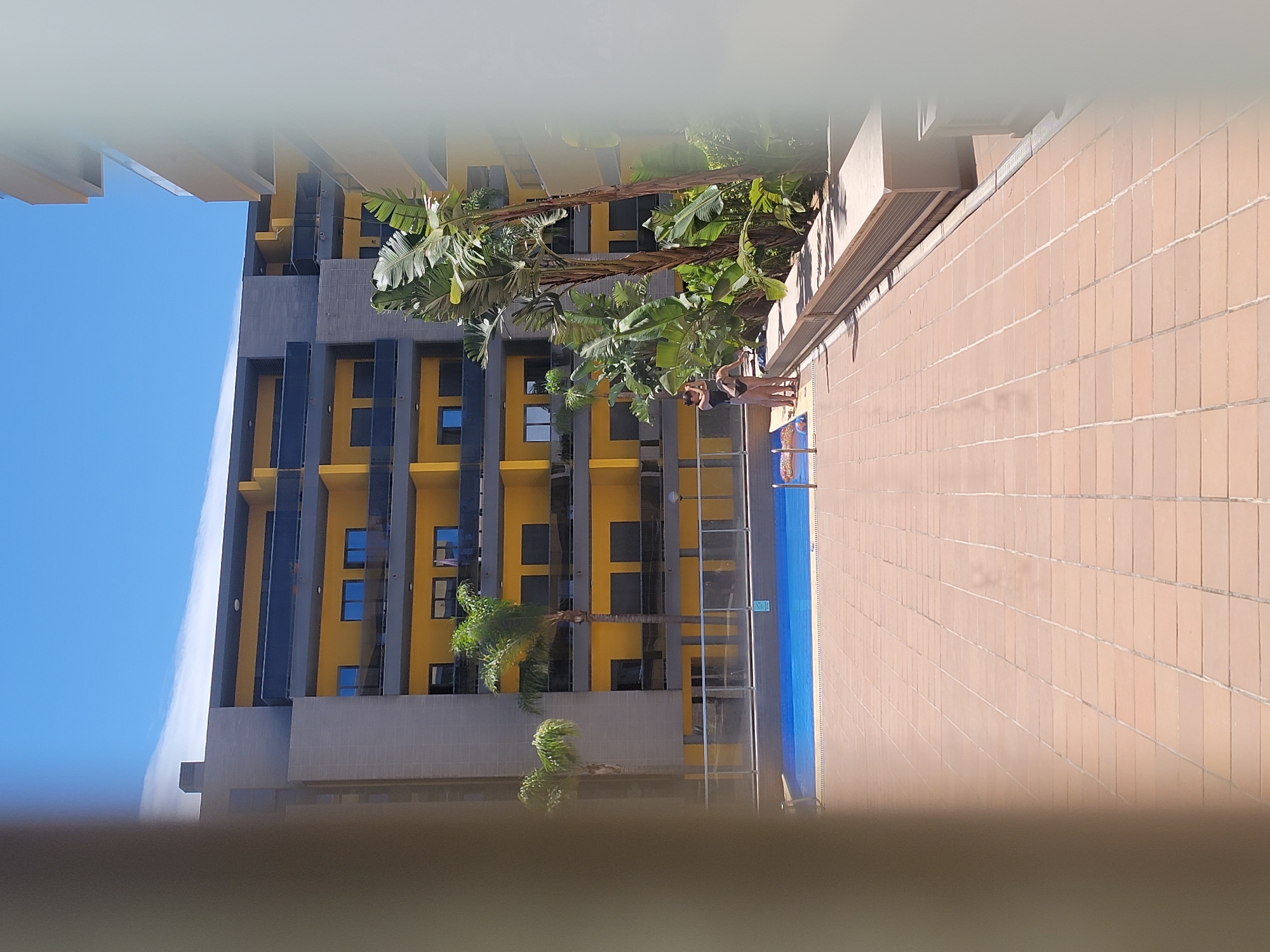 This is a medium shot of a residential building's courtyard in Quarteira, Portugal, on a bright, sunny day. The courtyard features a large blue swimming pool and a tiled patio area. Two women are standing near the edge of the pool, one in a black swimsuit and the other in a dark bikini. They appear to be looking towards the pool or at each other, with no clear interaction captured. The building itself is modern, with yellow facade, grey detailing, and dark balconies on multiple levels. Lush green plants, including palm trees and large-leafed tropical plants, are visible in the background and to the right, providing a tropical ambiance. The foreground is dominated by the tan tiled patio, with the edge of the pool visible to the left. The scene is framed by out-of-focus white and beige elements on the far left and right edges of the image, suggesting the photo was taken from a doorway or window.