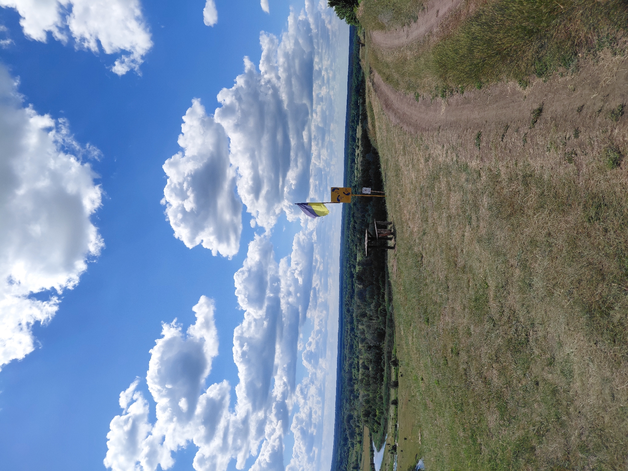 The image depicts a scenic view from a grassy hilltop overlooking a lush green valley under a bright blue sky dotted with fluffy white clouds. A dirt path winds its way up the hill, leading the viewer's eye towards the horizon. In the middle ground, a picnic table and bench are set up, suggesting a place for rest and contemplation. A Ukrainian flag flutters in the breeze, accompanied by a yellow sign with two figures dressed in what appear to be paragliding gear, indicating a potential paragliding spot. The valley below is densely forested, with a meandering river visible in the distance. The overall impression is one of natural beauty and outdoor recreation. The location is identified as Ustyvytsia, Ukraine.