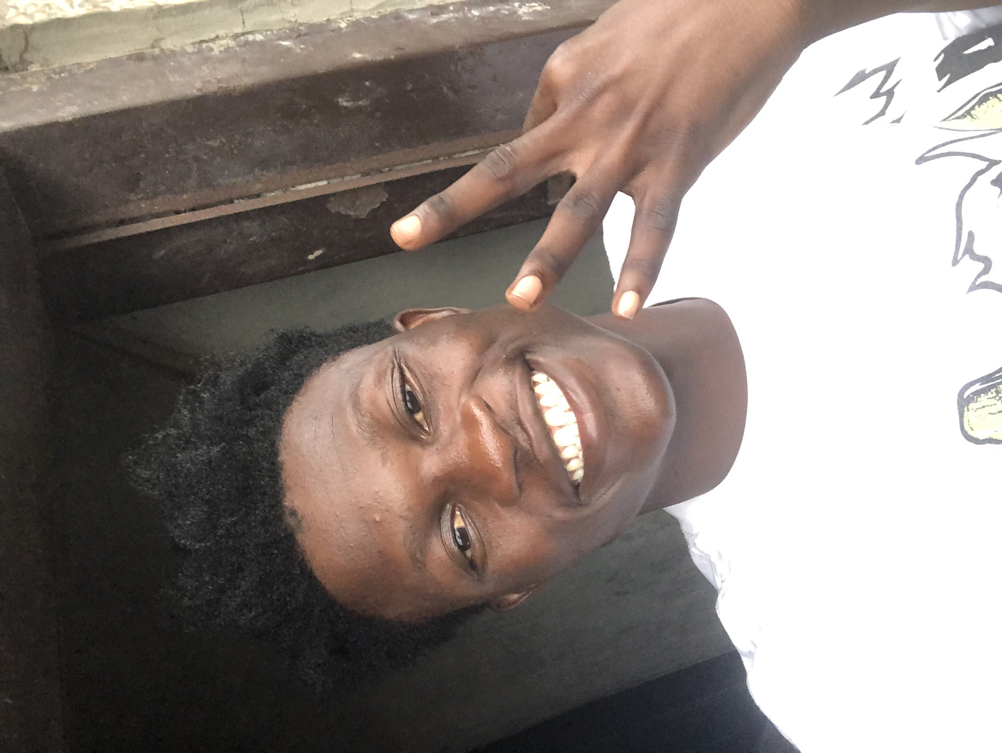 A young Black man with dark, curly hair smiles widely at the camera, showing off his teeth. He is wearing a white t-shirt with a graphic design partially visible on the front. His right hand is raised, with his index and middle fingers extended in a peace sign. The background appears to be an outdoor urban setting. The man's head and shoulders are the primary focus. The location is Ado, Nigeria.