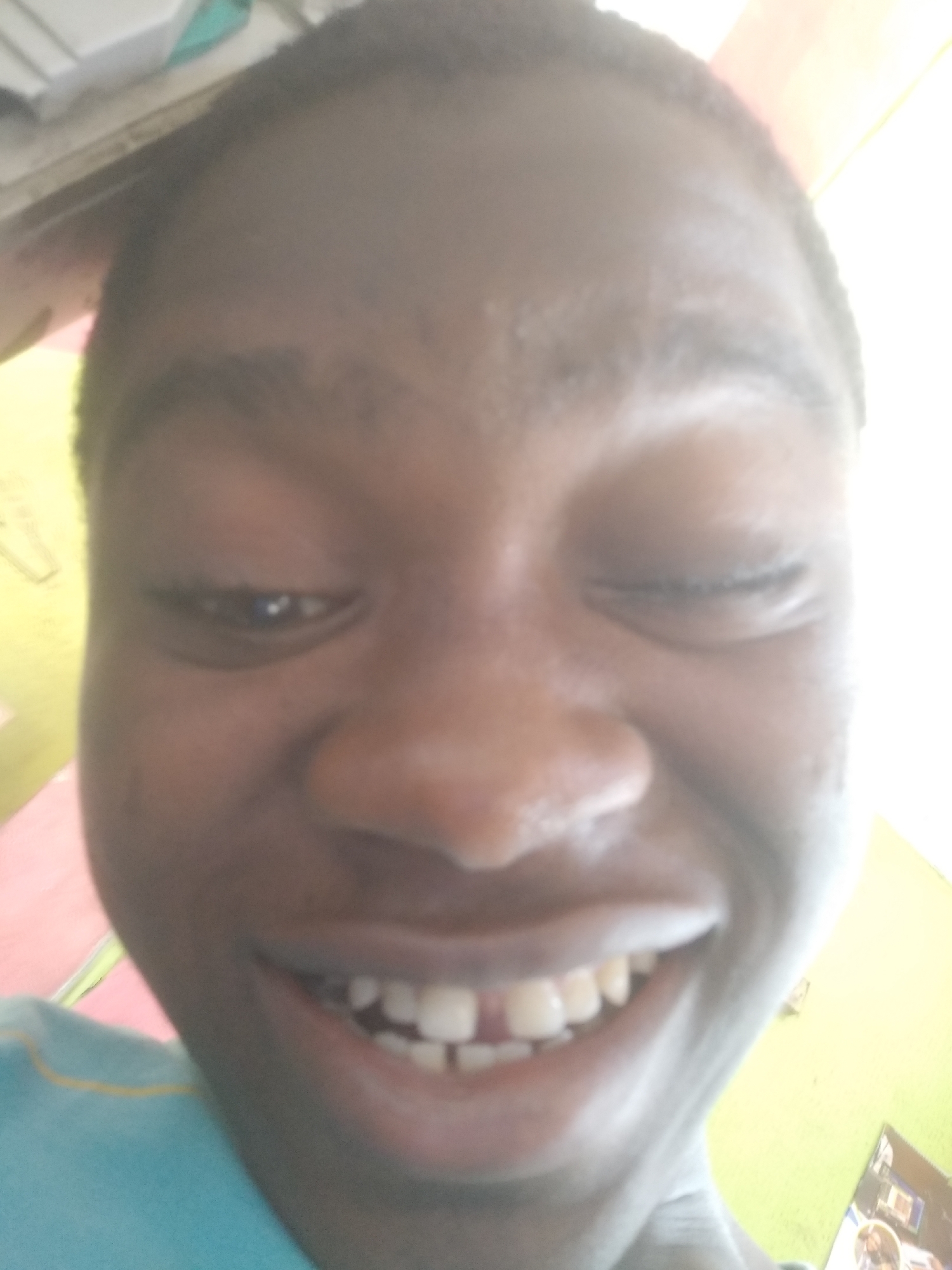A close-up selfie of a person with dark skin, one eye closed and a wide smile revealing teeth with a gap in the front. The person is wearing a blue shirt with a yellow stripe at the collar. The background includes a wall painted in shades of green, pink, and yellow. Part of a white cabinet is visible in the upper left. The presence of a poster with images suggests an interior space. The location is implied to be Shangisha, Nigeria.