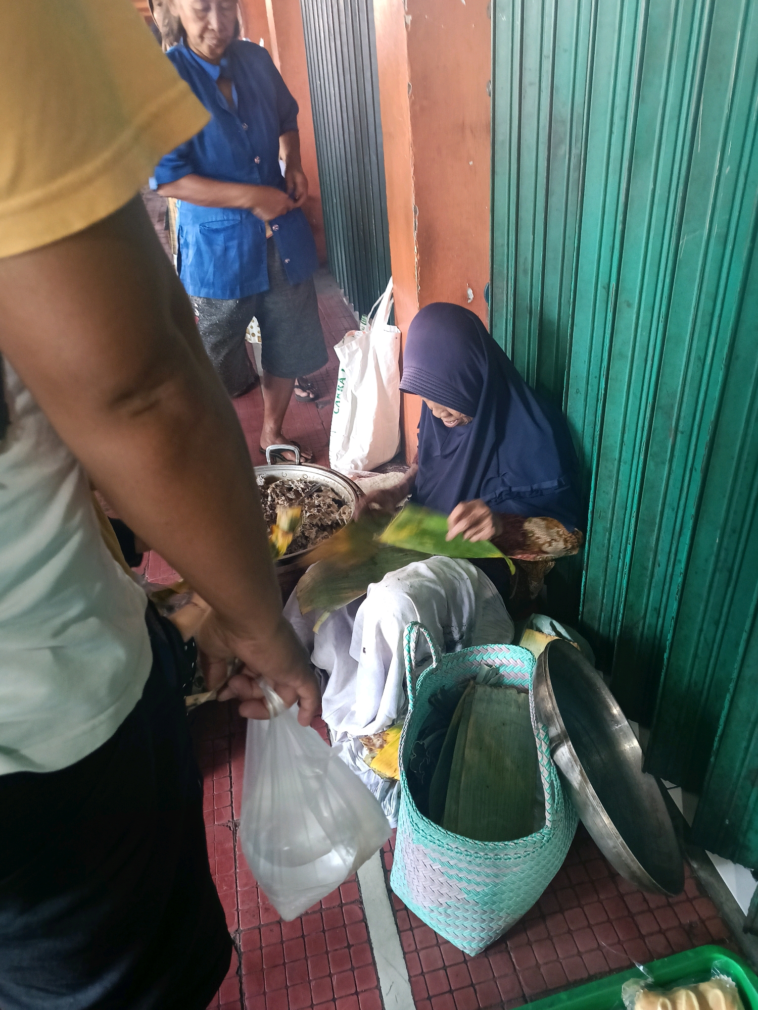 In Yogyakarta, Indonesia, a street vendor wearing a dark blue uniform and shorts is seen engaged in business. To her left, a person wearing a yellow shirt and dark pants is holding a transparent plastic bag filled with white objects. A woman in a dark blue hijab is seated on the ground, working with banana leaves. Beside her is a woven basket containing more banana leaves and a large metal pan rests on the ground. The background features a closed green metal shutter and a brown wall. The ground is tiled with red and white patterns.