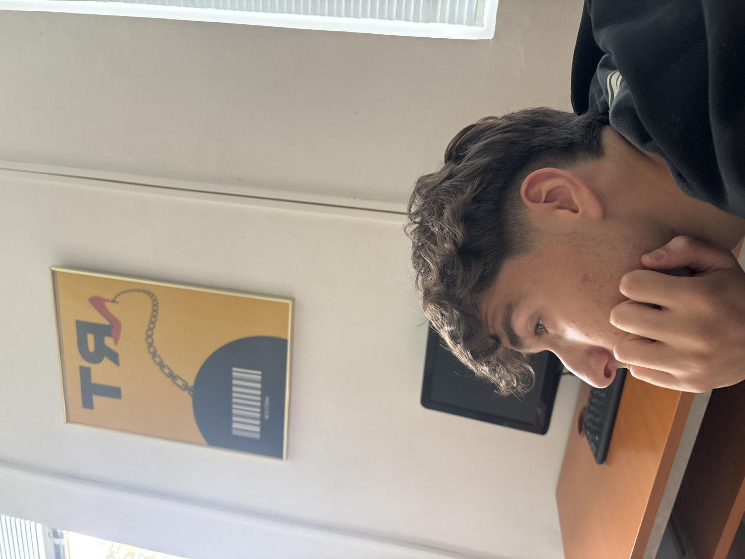 The image displays a young male in an indoor setting, likely a room in Troyan, Bulgaria, during the daytime. The individual, seen from a high angle and in profile, has short, curly dark hair and fair skin with some minor acne. He is wearing a dark hooded sweatshirt and is resting his left hand against his chin or cheek, looking downwards with a contemplative or focused expression.
Mounted on a light-colored wall behind him is a rectangular framed artwork or poster with a golden border. The poster features a vibrant yellow background. On the left, stylized text appears to be the Cyrillic letters "ТЯ" (which translates to "she"). A golden chain extends from the letter "Я" towards a large dark grey or black circular shape on the right side of the poster. Inside this circle, there is a white barcode pattern, and below it, faint, blurry white text that could be "HECTORY" or "НЕСТОРY".
Further to the right and slightly behind the person's head, a black electronic device, possibly a tablet or a small monitor, is visible. On a light brown wooden surface, presumably a desk, a black remote control or small keyboard is placed. The room is brightly lit, indicating natural light entering from a window or light source, partially visible with blinds in the upper and lower left corners. The scene suggests a calm and quiet atmosphere, with the individual appearing to be in thought or focused on something out of frame.