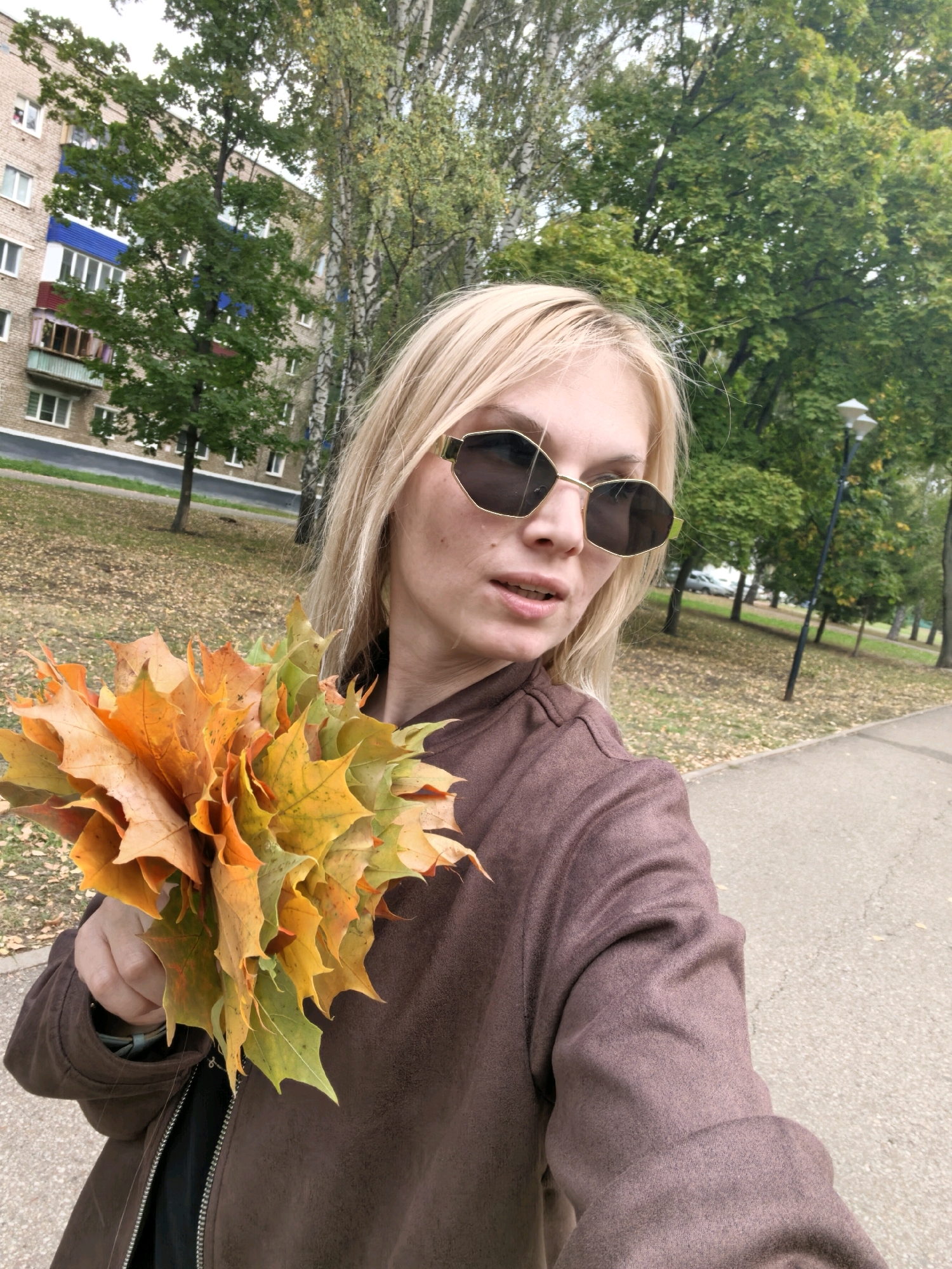 A woman with blonde hair and sunglasses is holding a bouquet of colorful autumn leaves. She is standing on a paved path in an outdoor setting. In the background, there is a brick apartment building and trees with green and yellow leaves. The ground is covered with fallen leaves. The overall scene suggests an autumn day in an urban park or residential area.