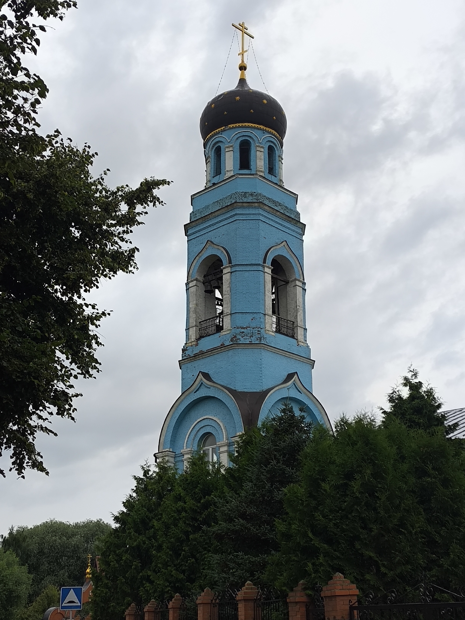 The image shows a blue brick bell tower of a church with a black dome topped by a golden cross. The tower has arched openings where bells are visible. Dense green trees partially obscure the lower portion of the tower. The sky is overcast and cloudy. A traffic sign with a triangle and a hill symbol is visible in the lower left corner of the image, and a section of a brick fence is in the foreground. This structure is part of the Holy Trinity Convent located in the city of Pokrov, Russia.