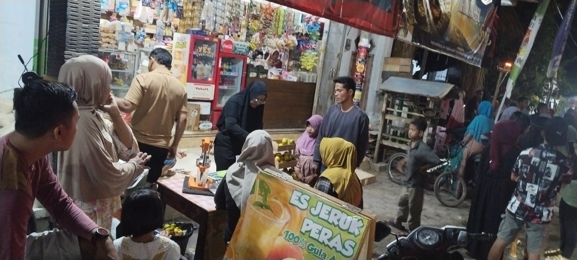 The image depicts a street scene in Keting, Indonesia, likely a bustling marketplace or a collection of small shops. In the foreground, a man in a maroon shirt is leaning forward, looking to his left, with a watch on his left wrist. To his right, a child with dark hair and wearing a white shirt is visible. Behind the man in the maroon shirt, a woman in a light brown hijab is talking, and a man in a tan shirt is looking away.
Further into the scene, a woman in a black hijab is attending to something on a table that holds a manual juice press. A child in a purple hijab is standing nearby. A sign with "ES JERUK PERAS 100% GULA A" is prominently displayed in the foreground. In the background, shelves are stocked with various items, and a red refrigerator with "YES EVERYDAY" and Coca-Cola branding is visible. Other individuals are present, some wearing hijabs of various colors, contributing to the crowded atmosphere. A bicycle is parked near a shelf unit on the right side of the image, and a motorbike is parked in the lower right corner. The environment appears to be outdoors, illuminated by artificial lighting.