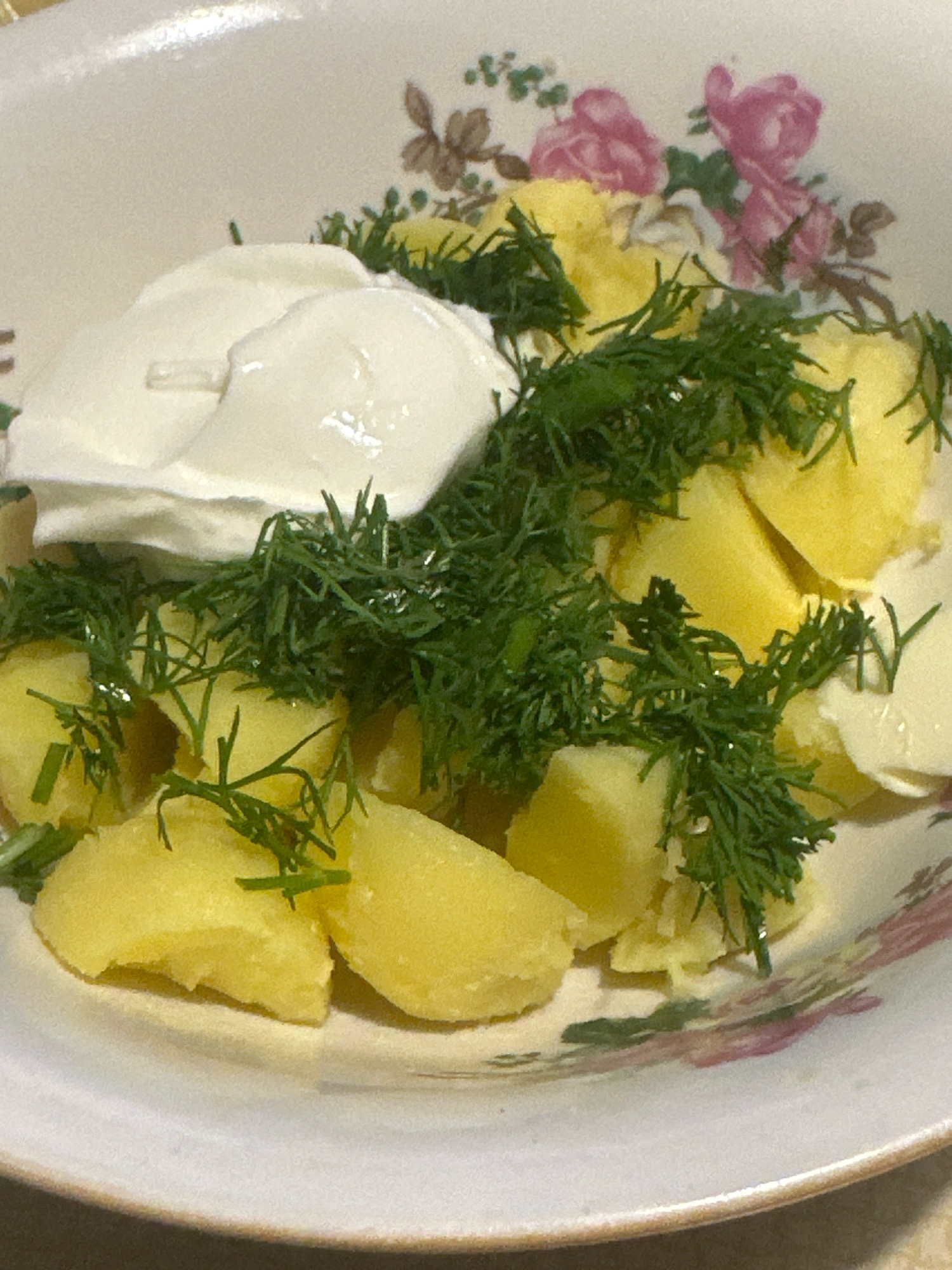 A close-up, overhead shot captures a serving of boiled potatoes in a white bowl with a floral pattern. The potatoes are cut into bite-sized pieces and are topped with a dollop of white sour cream or yogurt and a generous sprig of fresh green dill. The bowl is decorated with pink and brown floral motifs. The background is blurred and suggests a neutral surface, possibly a table. No people are visible in the image. The scene is domestic and appears to be a simple, home-cooked meal. The location in Biysk, Russia, is not visually evident from the image itself.