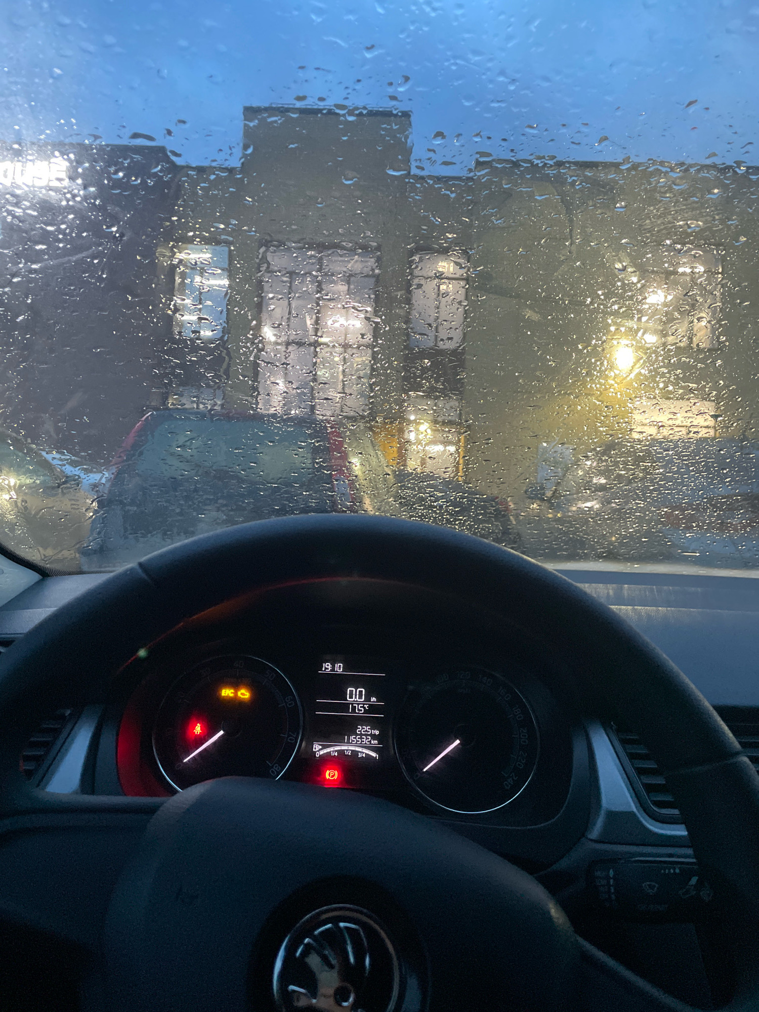 The image, captured from the interior of a vehicle, shows a view through a rain-streaked windshield. Outside, a building with multiple windows is visible, illuminated from within. The time displayed on the car's dashboard is 19:10, and the temperature is 17.5°C. Several warning lights are illuminated on the dashboard, including an EPC (Electronic Power Control) light and a seatbelt reminder. The car's odometer shows 115532 km. The scene is consistent with an evening drive in Arkhangelsk, Russia, during rainy weather.