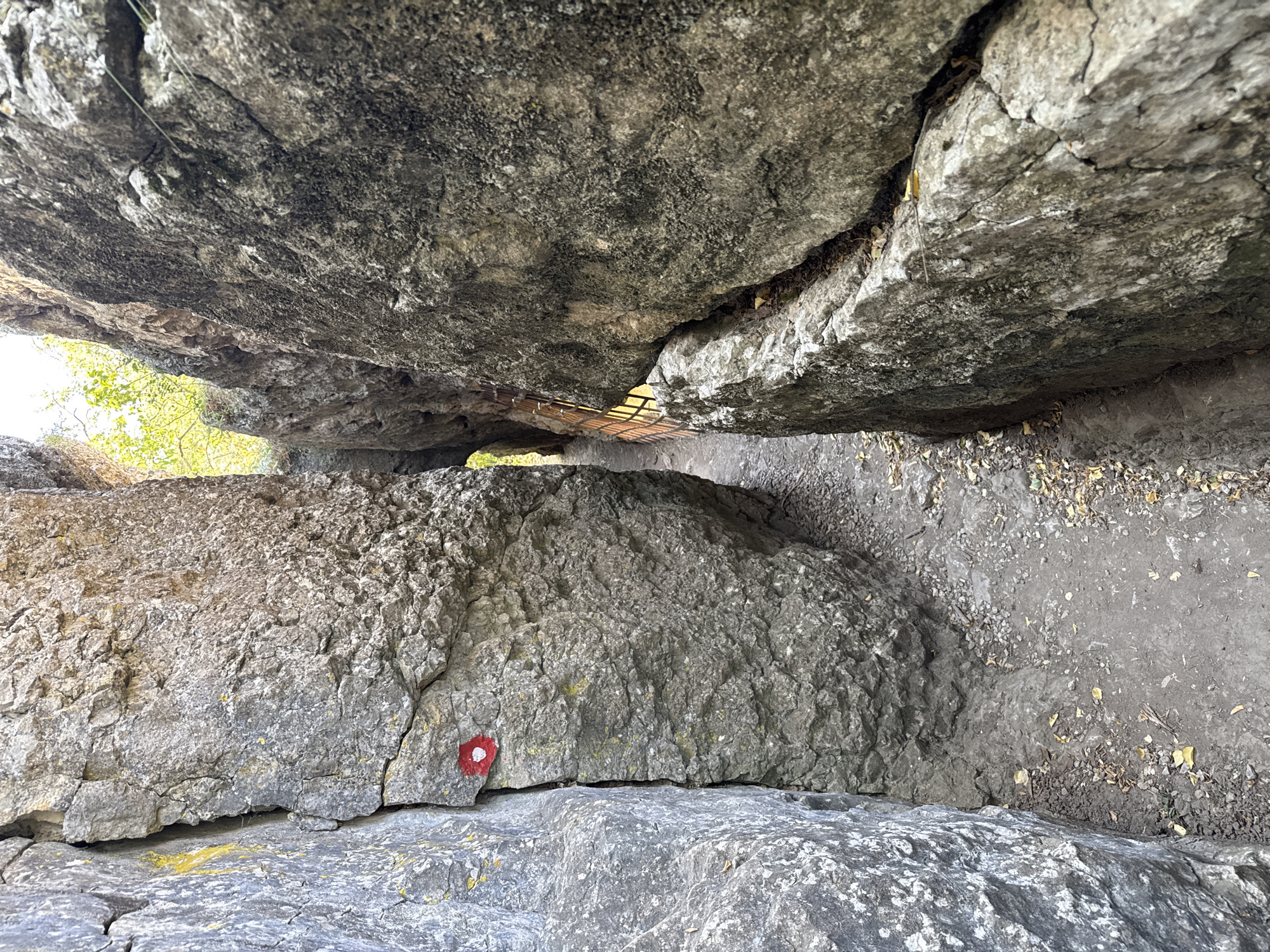 The image shows a narrow path between two large, rough rock formations. The path is covered with dirt and small stones, with some fallen leaves scattered about. The rock on the left has a red and white circular marking on it, possibly a trail marker. The rock on the right is darker and more textured. In the distance, the path appears to open up to a brighter area with foliage visible. The scene depicts a natural landscape, likely a hiking trail within a rocky canyon. This specific location is part of the Hotnitsa Waterfalls area in Bulgaria, known for its natural beauty and hiking opportunities.